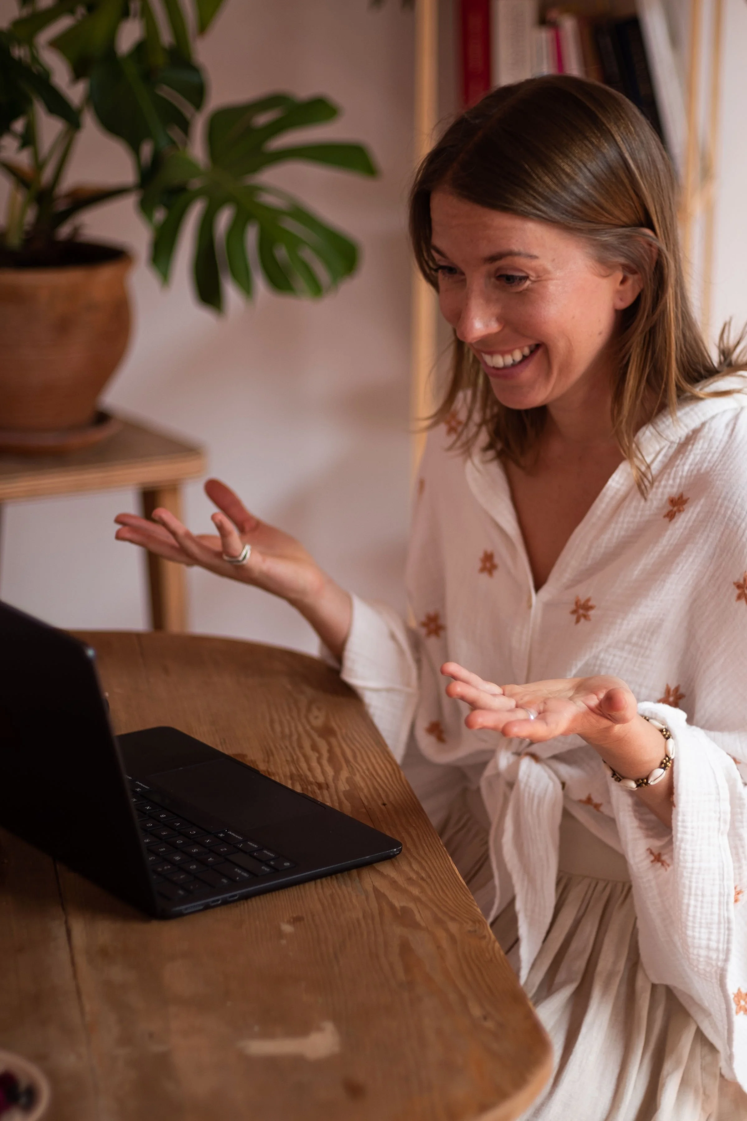 A woman with shoulder-length light brown hair smiling and gesturing with her hands while sitting at a wooden table with a black laptop in front of her. She is wearing a white blouse with orange floral embroidery and a beaded bracelet.