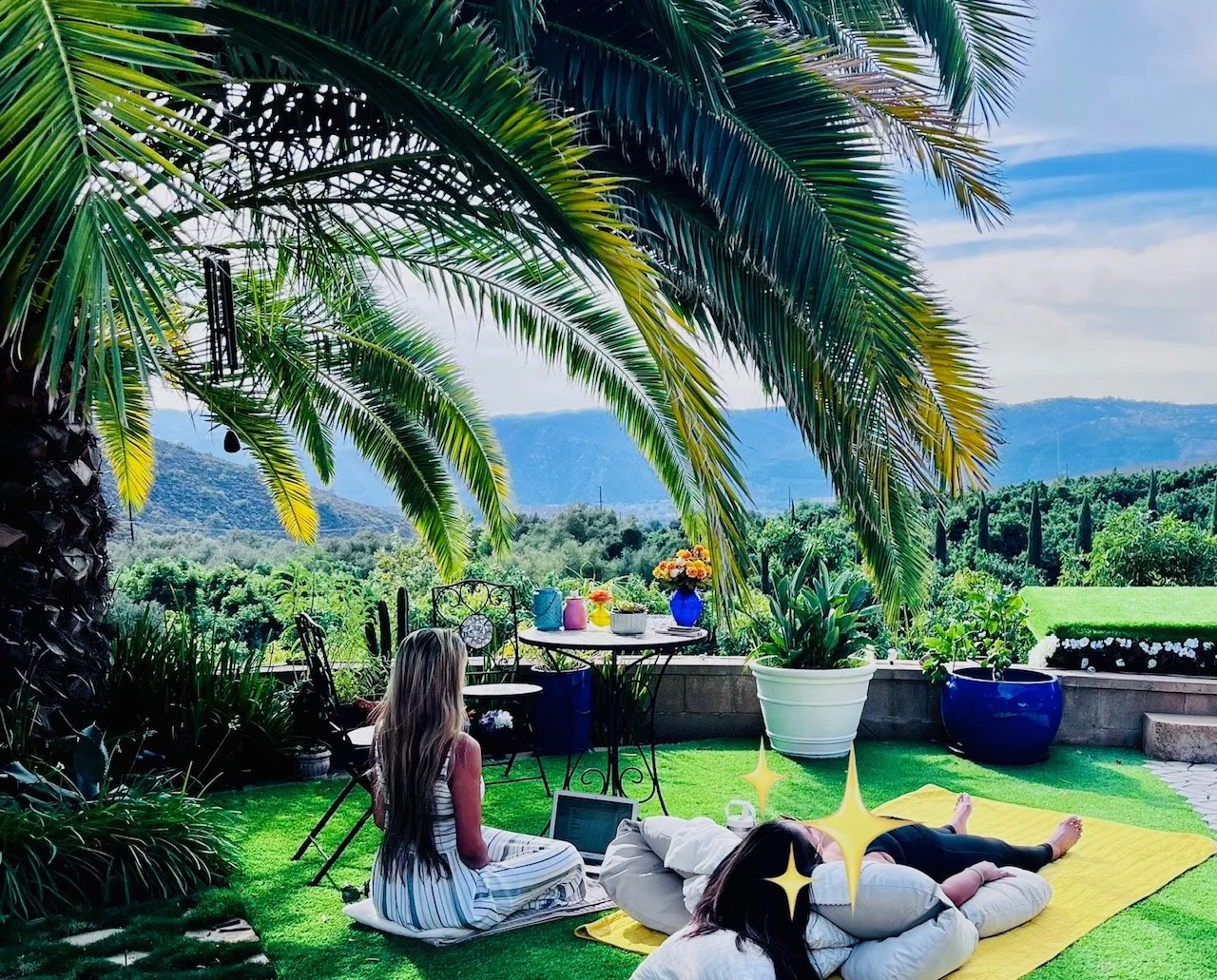 People relaxing on a rooftop garden with lush greenery, palm trees, and mountains in the background. One person is seated using a laptop, while another is lying on a yellow blanket. There is a table with colorful vases and flowers, and large potted plants surround the area.