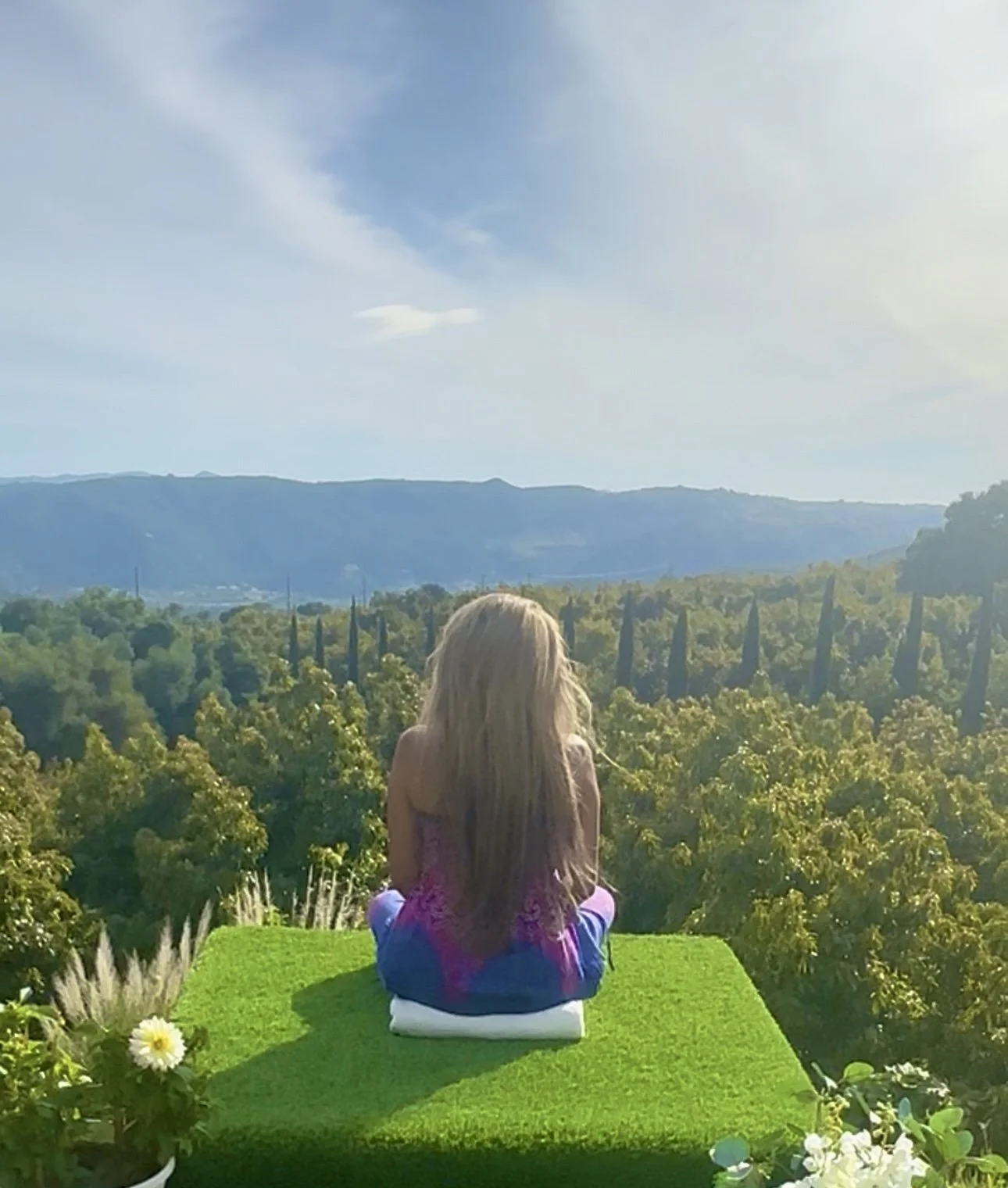 A young girl with long blonde hair is sitting cross-legged on a green mat, facing away, in a garden with yellow flowers, overlooking a landscape of green trees, rolling hills, and blue mountains under a partly cloudy sky.