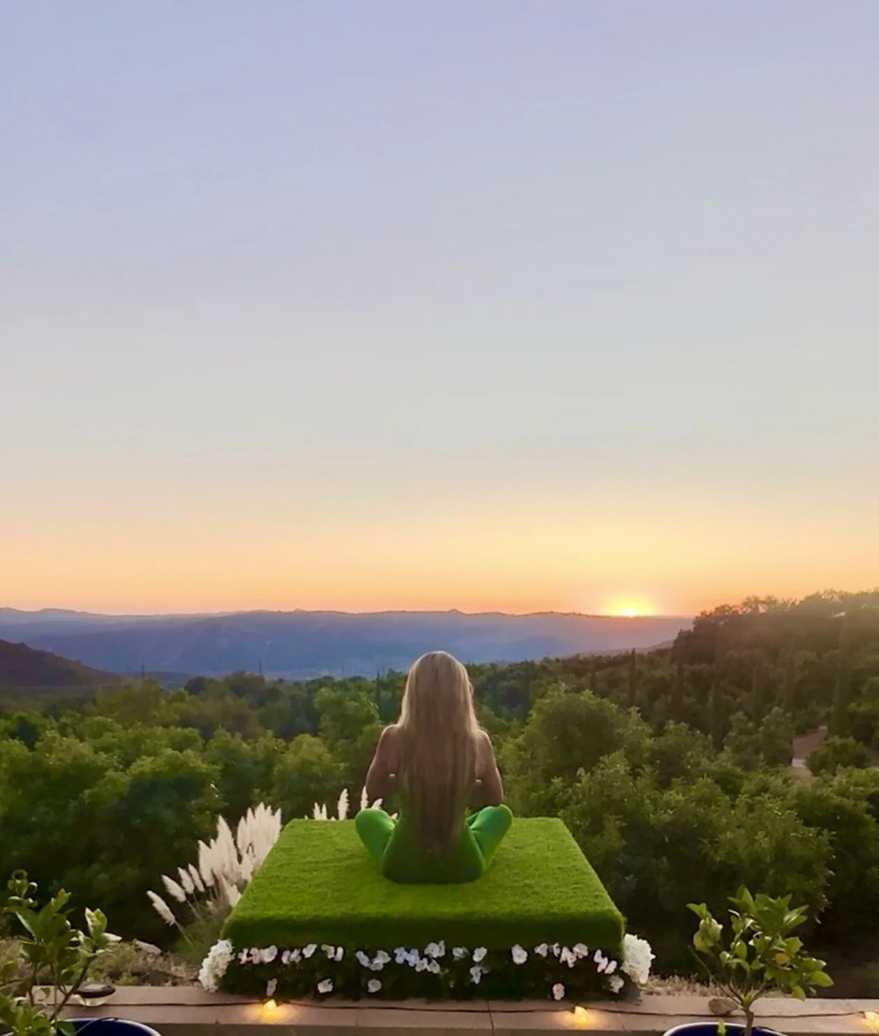 A woman with long blonde hair sits cross-legged on a grassy platform, facing a sunset over mountains and trees, with decorative plants and lights around her.