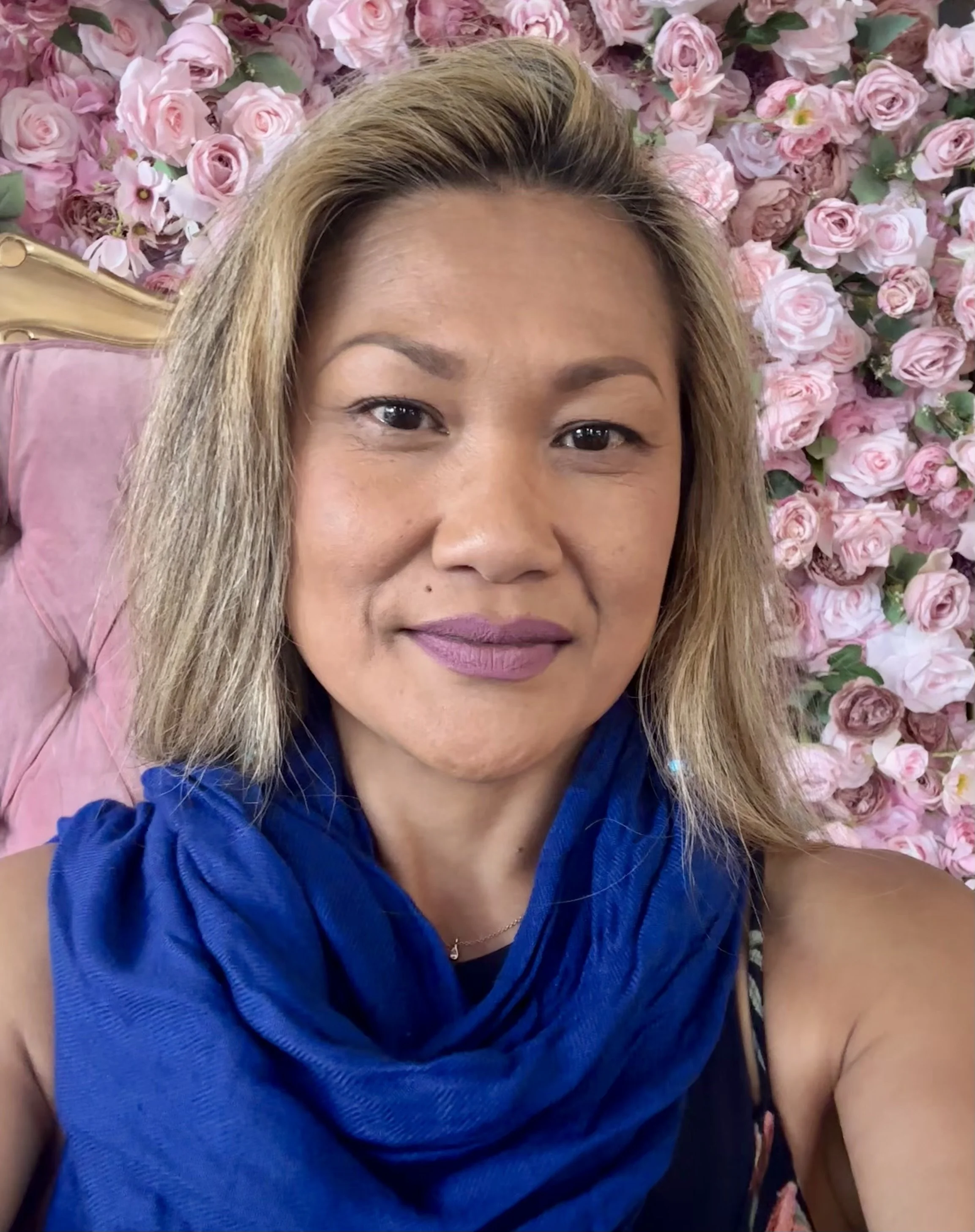 A woman with light brown hair and a floral-patterned top taking a selfie in a museum or gallery with classical pottery and artworks displayed in the background.