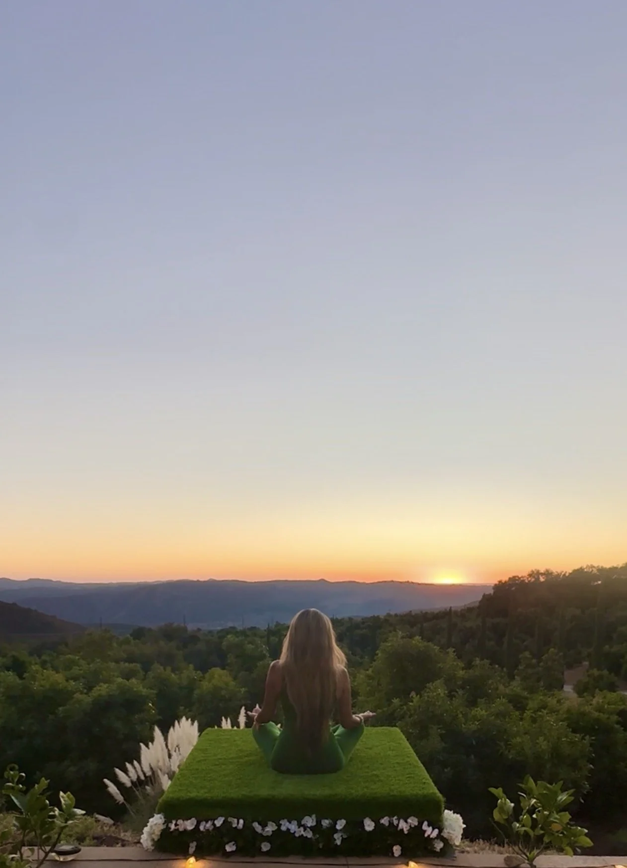 A young girl with long blonde hair sitting on a green mat facing a scenic view of rolling hills and blue sky, surrounded by flowering plants.