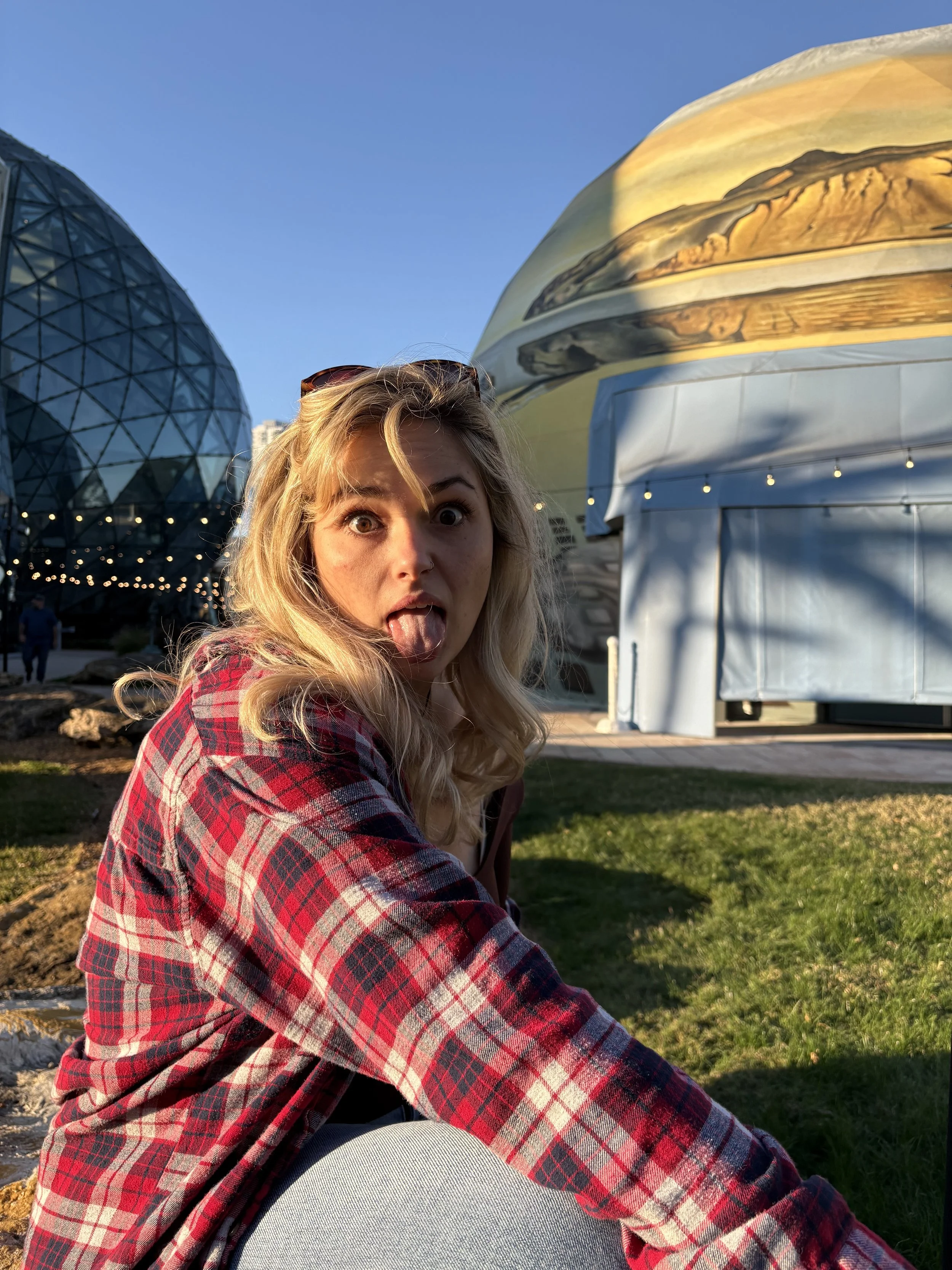 A woman with blonde hair making a silly face with her tongue out, wearing a red and black plaid shirt and sunglasses on her head, sitting outdoors with modern buildings in the background during sunset.