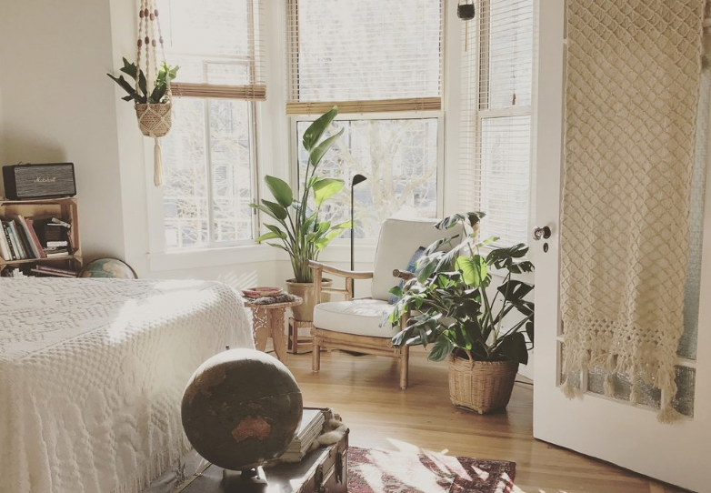 Sunlit bedroom with a bed, a globe on a small table, green houseplants, a white chair, and a door with a lace curtain.