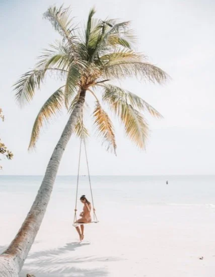 A girl sitting on a swing hanging from a palm tree on a sandy beach with calm ocean in the background.