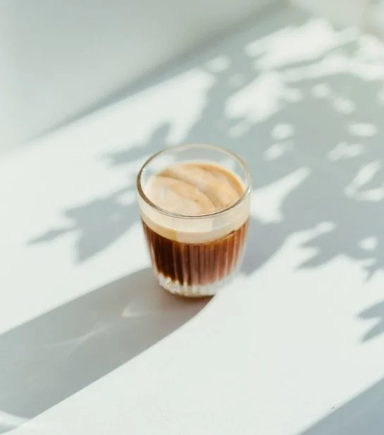 A glass of frothy iced coffee on a white surface with shadows of leaves.