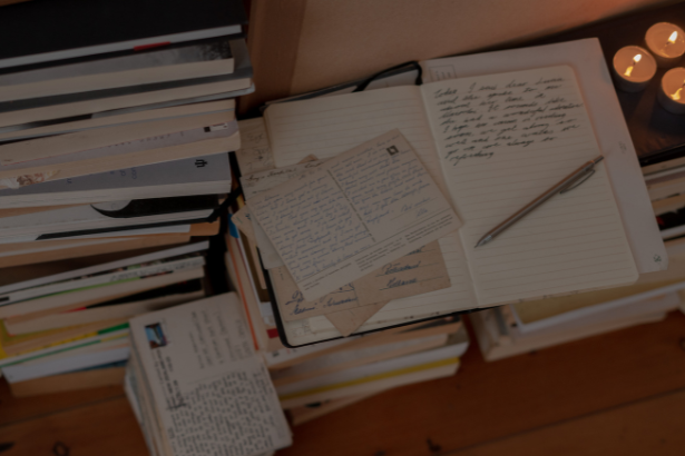 A cluttered desk with open notebooks, handwritten notes, and postcards, surrounded by stacks of books and candles.