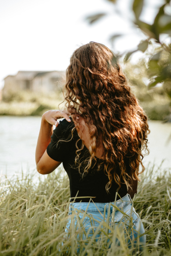 A woman with long, curly hair sitting outdoors near a body of water, surrounded by grass and trees, with her back to the camera.
