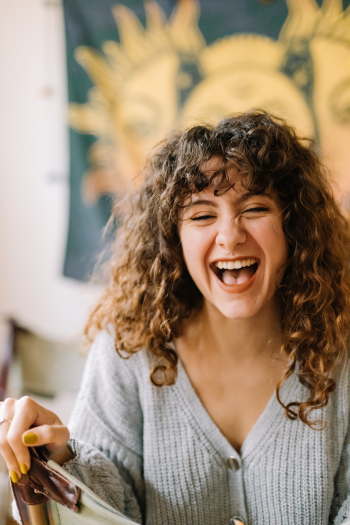 A young woman with curly hair smiling and laughing while holding a pen, with a colorful wall art featuring sun and moon behind her.