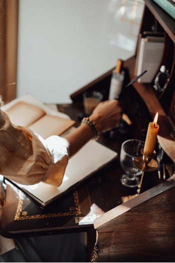 Person writing in a notebook at a wooden desk with a candle, glass of water, and books.