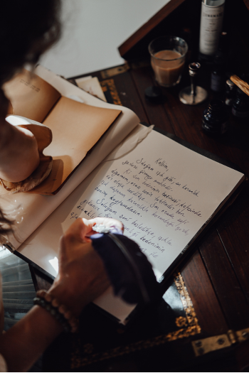 A person writing in a notebook with a pen and an open book nearby on a wooden desk. There is a glass of water and some bottles on the desk.