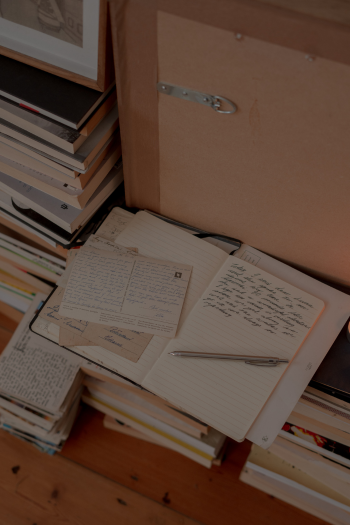 Open notebooks with handwritten notes and loose papers on a desk, surrounded by stacks of books.