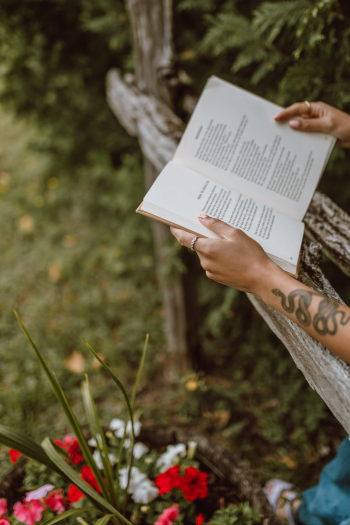 Person reading a book outdoors near a tree and flowers.