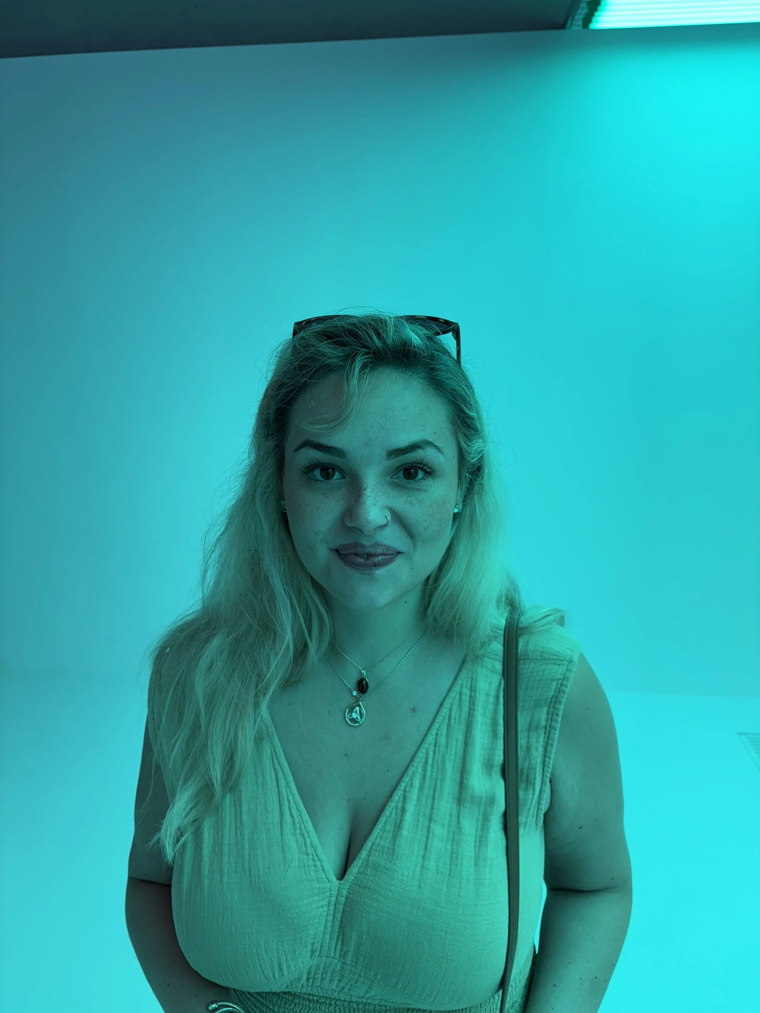A woman with blonde hair, blue eyes, and freckles wearing a yellow top and two necklaces, standing against a blue and teal background with teal lighting.