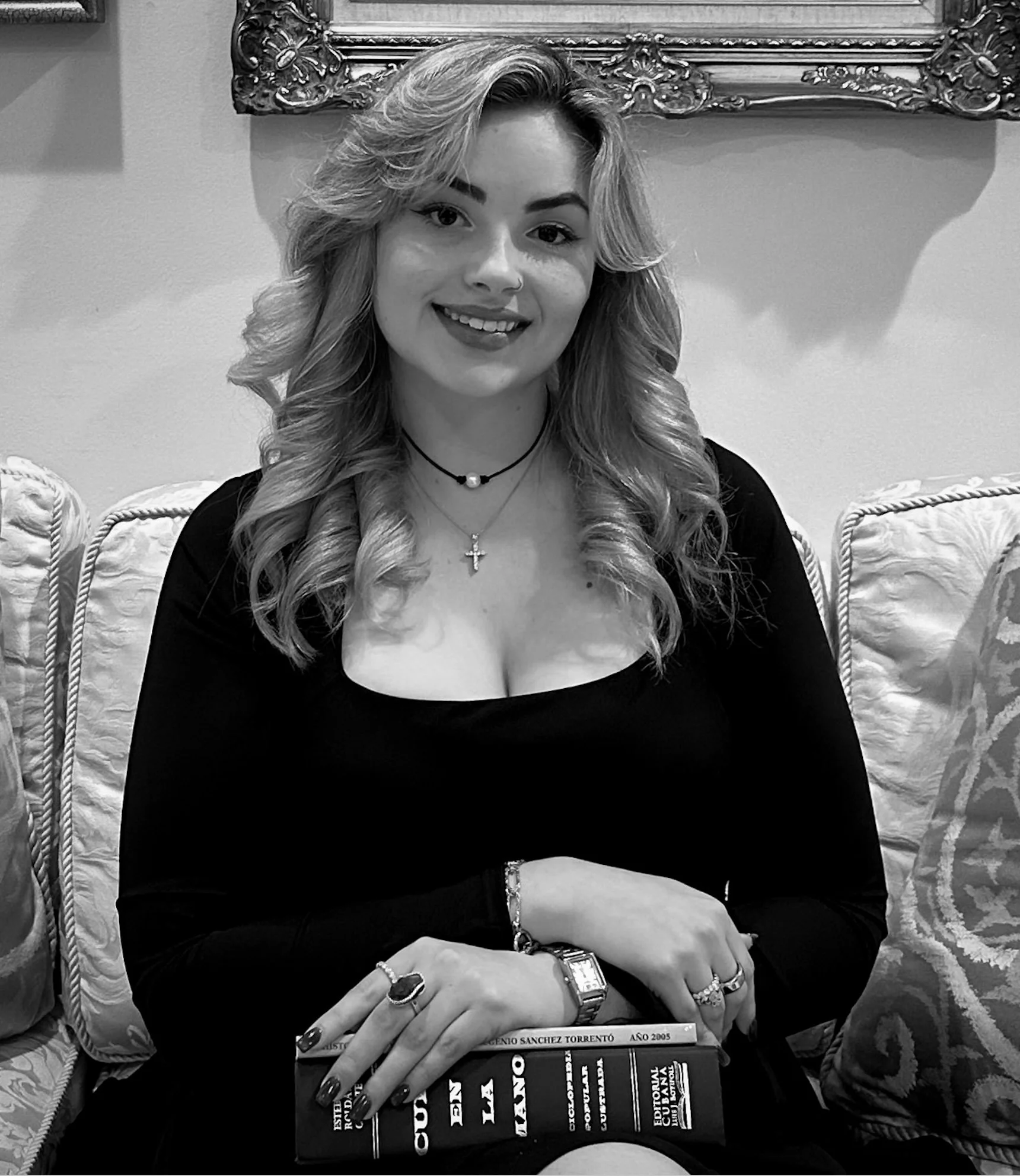 A woman with wavy hair, wearing a black top, silver jewelry, and a cross necklace, sitting on an ornate chair, holding a book on her lap, with patterned pillows and a large decorative mirror in the background.