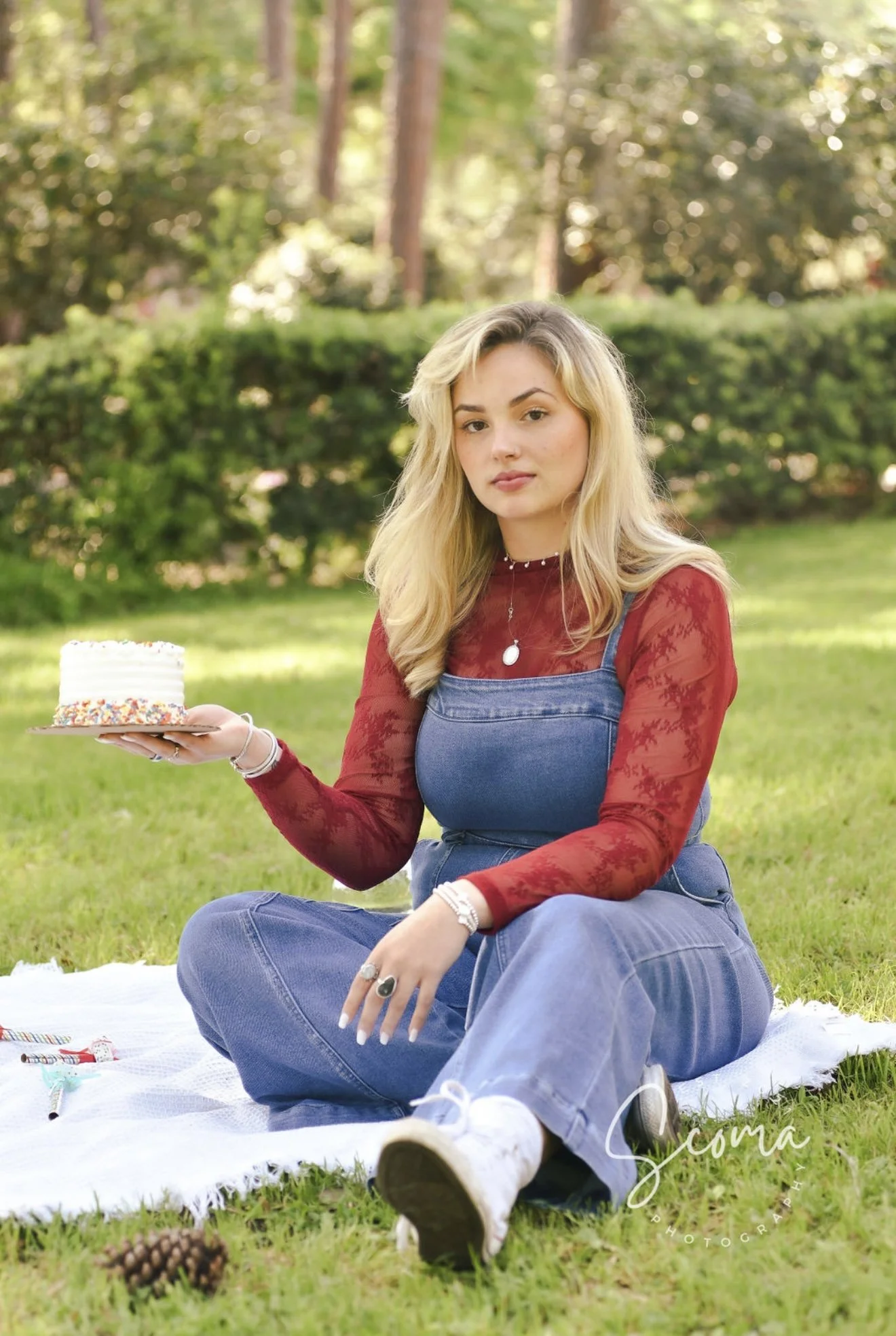 A young woman with blonde hair and fair skin sitting on a white blanket in a park, holding a small birthday cake decorated with colorful sprinkles. She is wearing a red long-sleeve top and blue overalls. The background features green trees and bushes, with sunlight filtering through.
