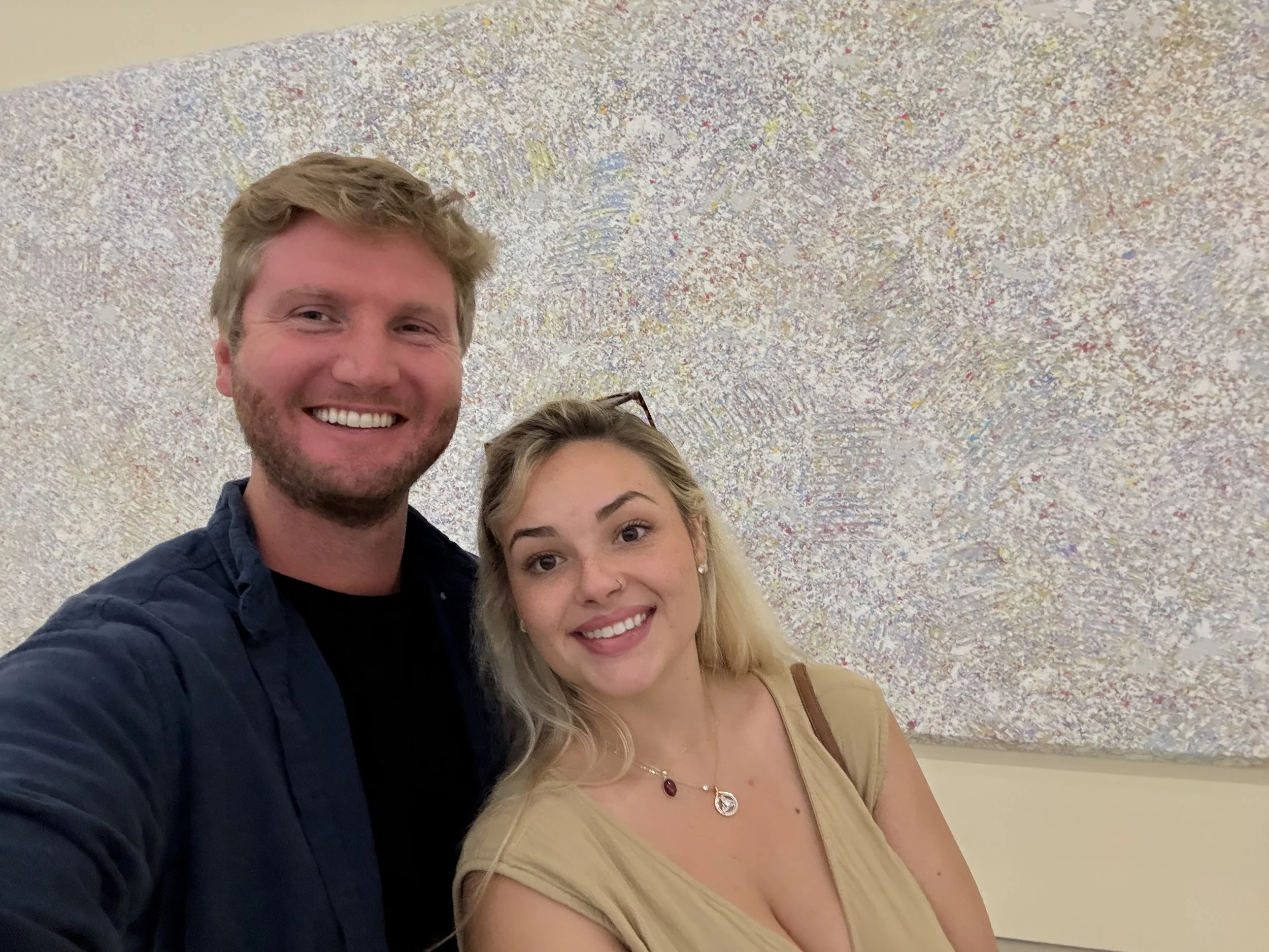 A smiling man with light brown hair and a beard taking a selfie with a smiling woman with blonde hair and a nose ring, standing in front of a colorful abstract painting.