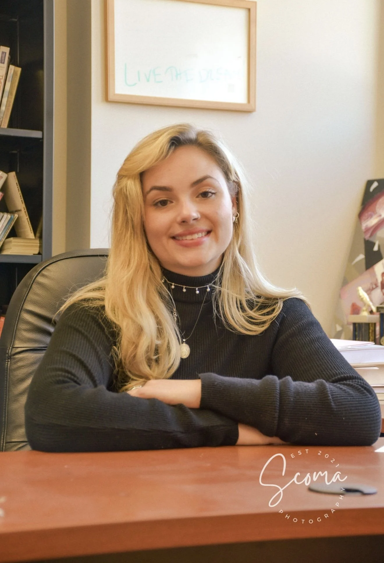 A young woman with long blonde hair, wearing a black turtleneck sweater and jewelry, sitting at a wooden desk in an office. Behind her are bookshelves and a whiteboard with writing on it.