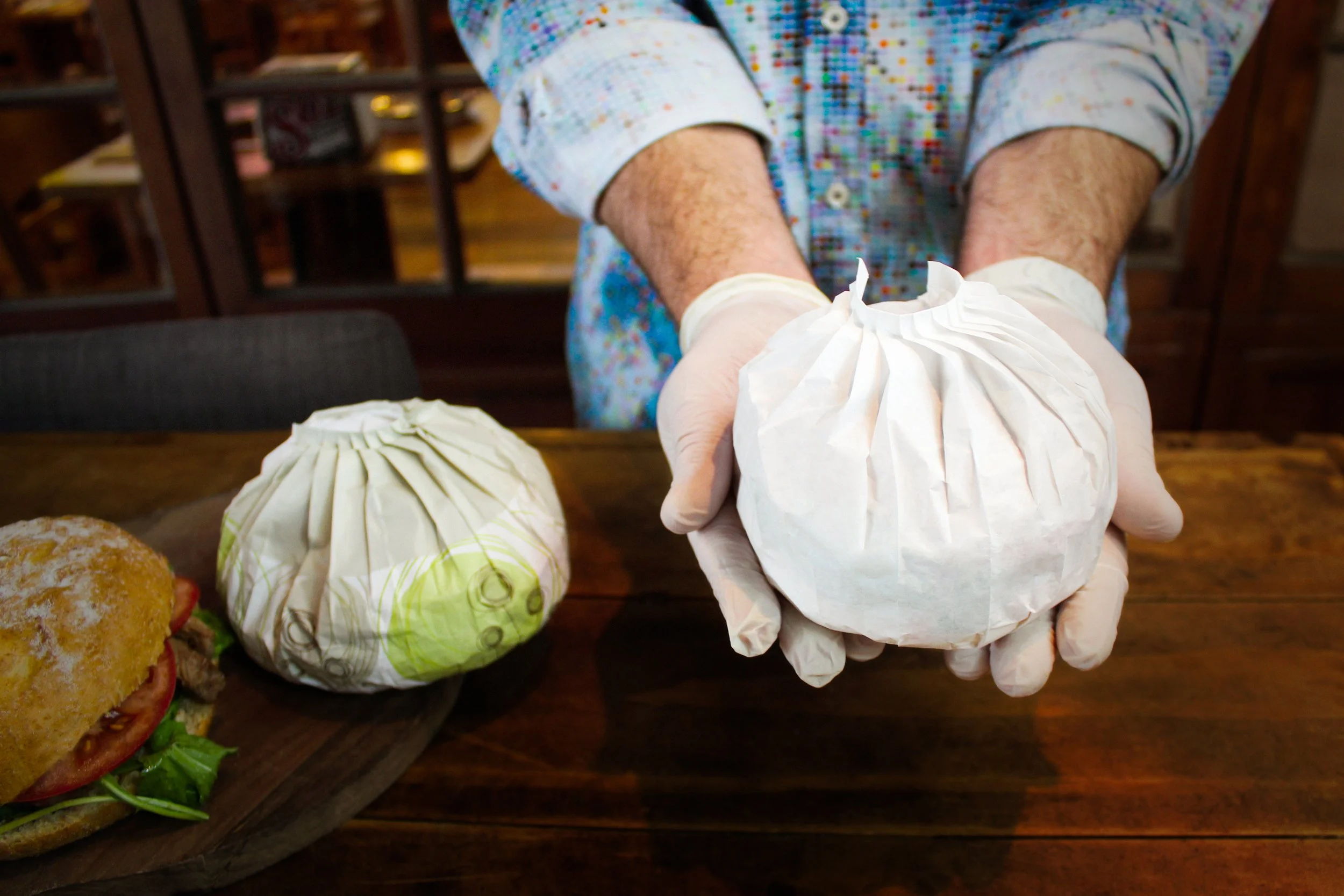 Person wearing white gloves holding a burger wrapped in white paper wrapping.