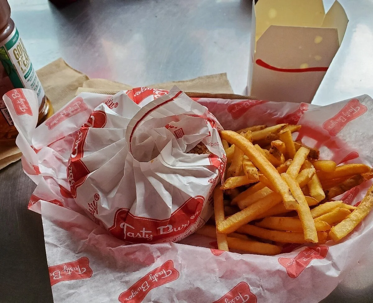 A burger wrapped in red and white paper, a side of French fries, and a small container of dipping sauce on a paper-lined tray at a fast-food restaurant.