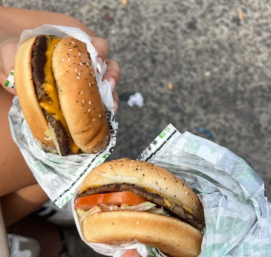 Two cheeseburgers, one with tomato, lettuce, and cheese, held in hand with paper wrapping, outdoors on a gray surface.
