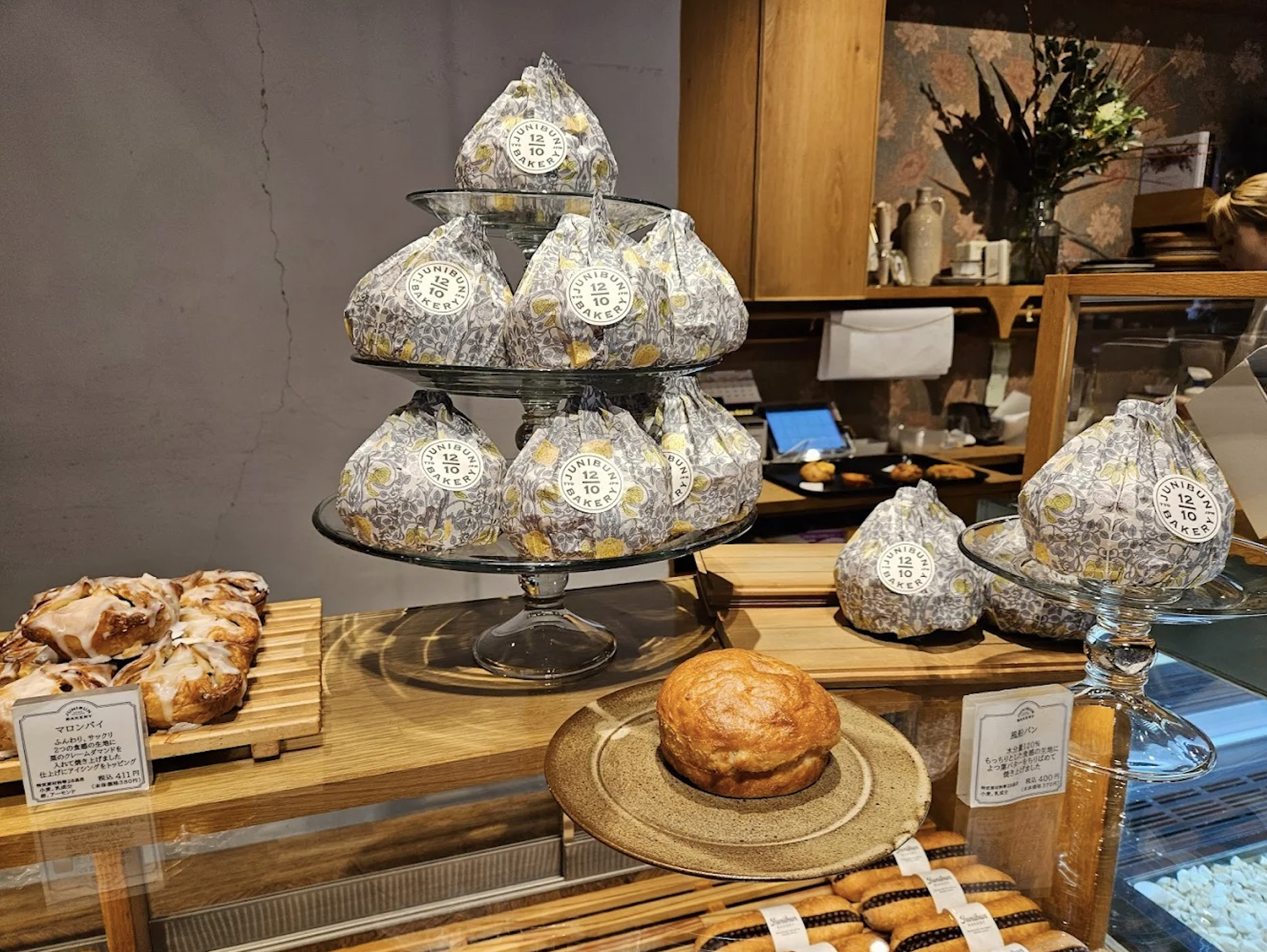 Display case with wrapped baked goods on a tiered stand and a single bread on a plate, inside a bakery shop.