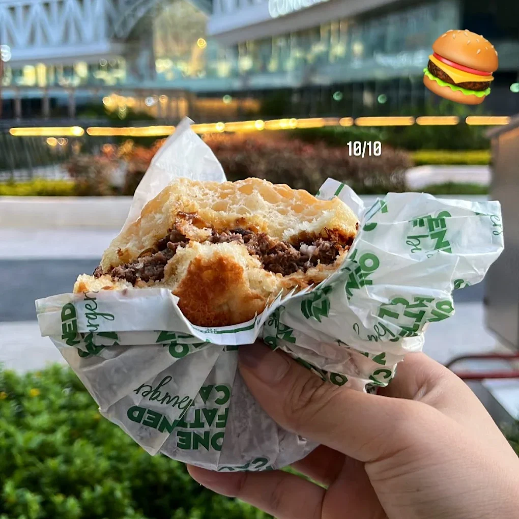 Close-up of a person holding a half-eaten cheeseburger with lettuce, tomato, and a beef patty in white paper wrap, outdoors with blurred plants and a walkway in the background.