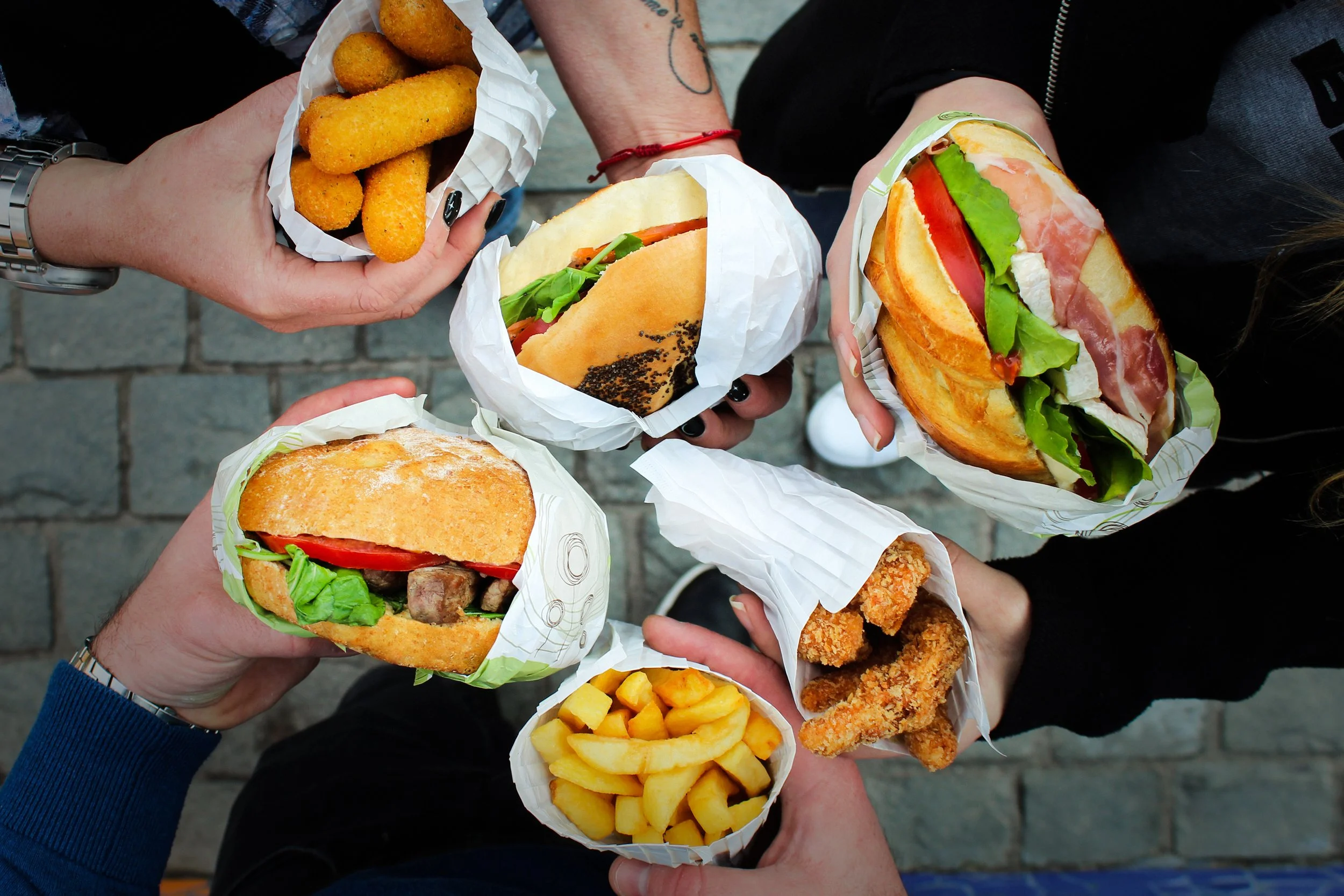 Overhead view of four hands holding burgers, fries, and fried foods, with a cobblestone surface underneath.