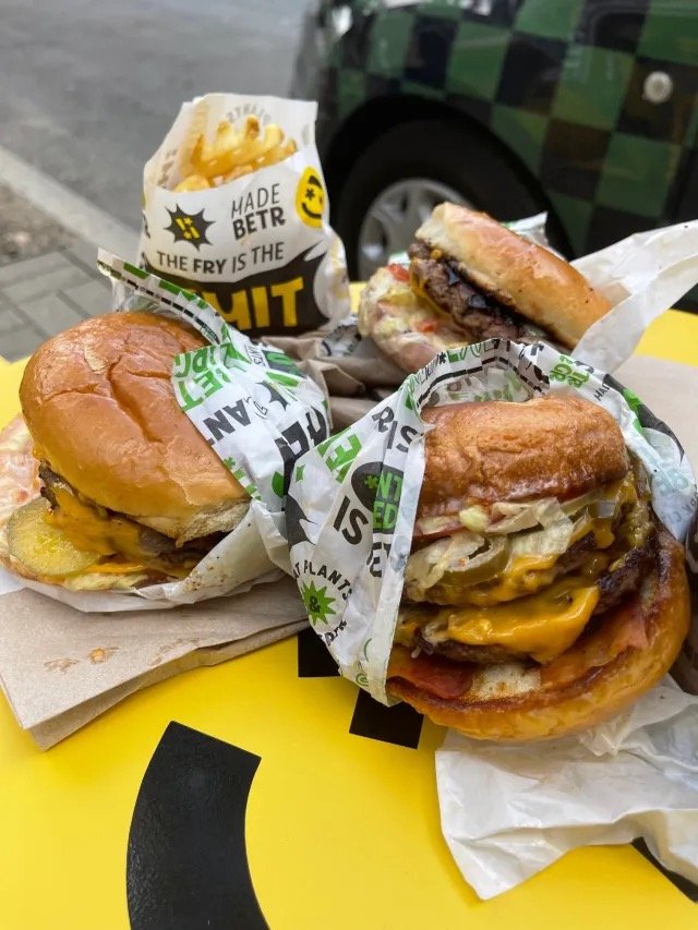 Three cheeseburgers and a cup of french fries on a yellow table outdoors.