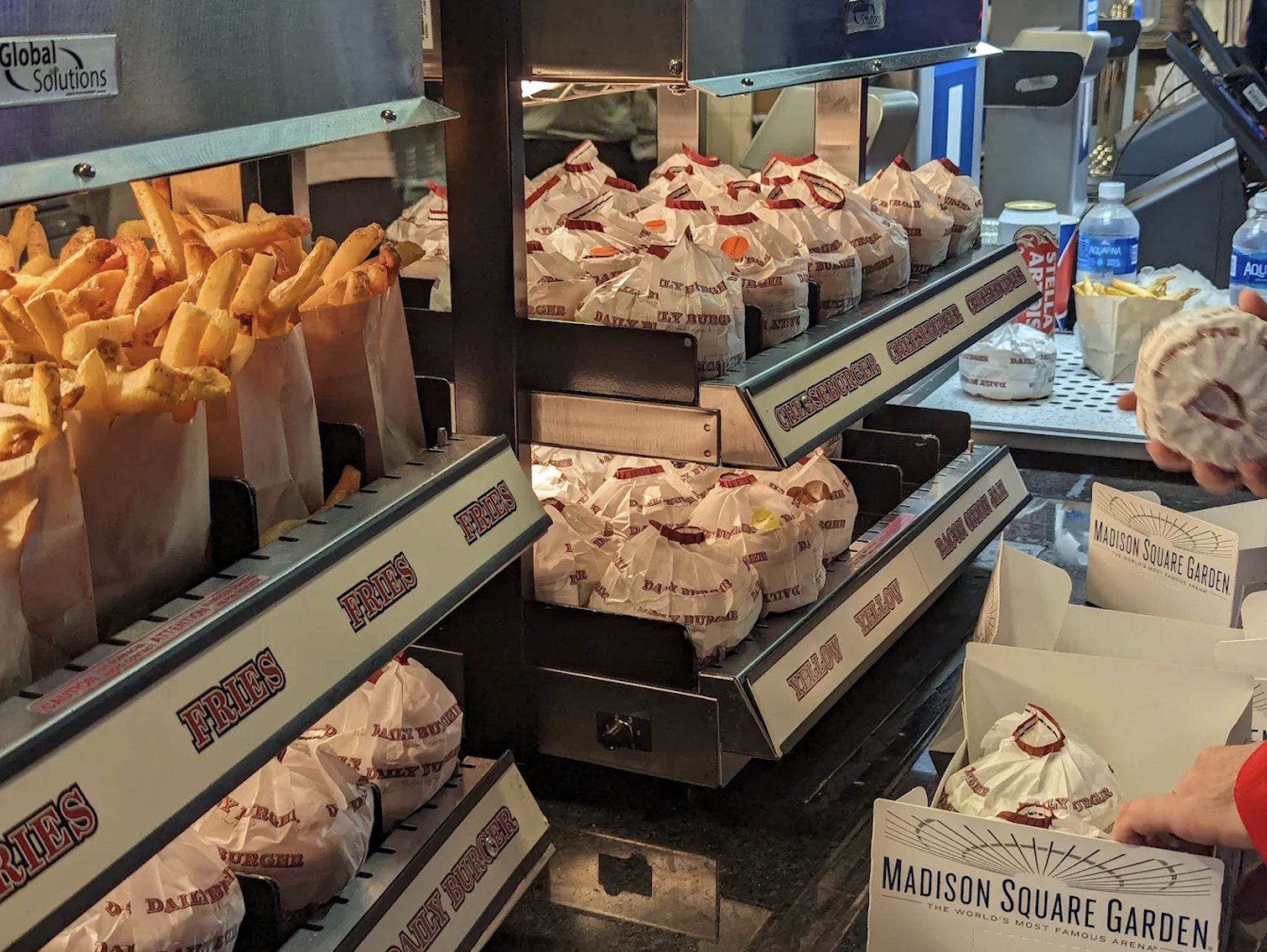 Fast food restaurant counter with paper-wrapped burgers, baskets of French fries, and soft drinks on display.