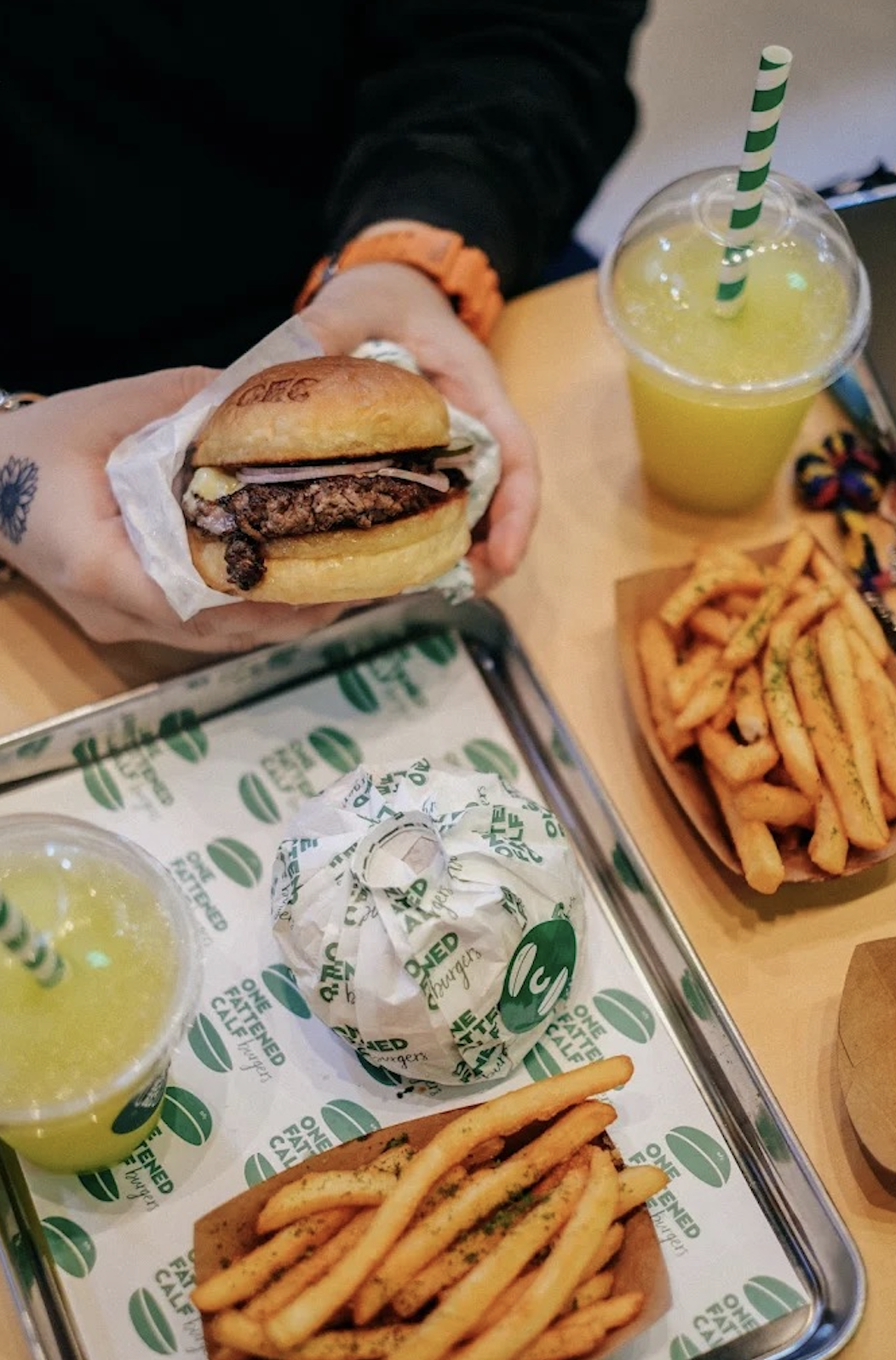 Person holding cheeseburger with onions, fries, and drinks on tray at a restaurant.