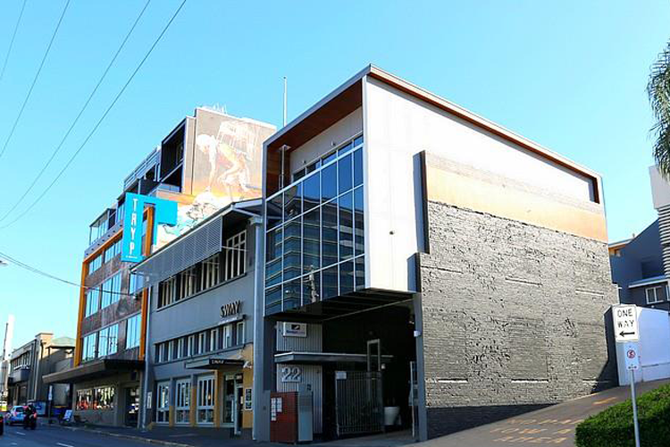 Modern multi-story building with glass windows and signage for a hotel, situated on a city street with parked cars and a one-way street sign.