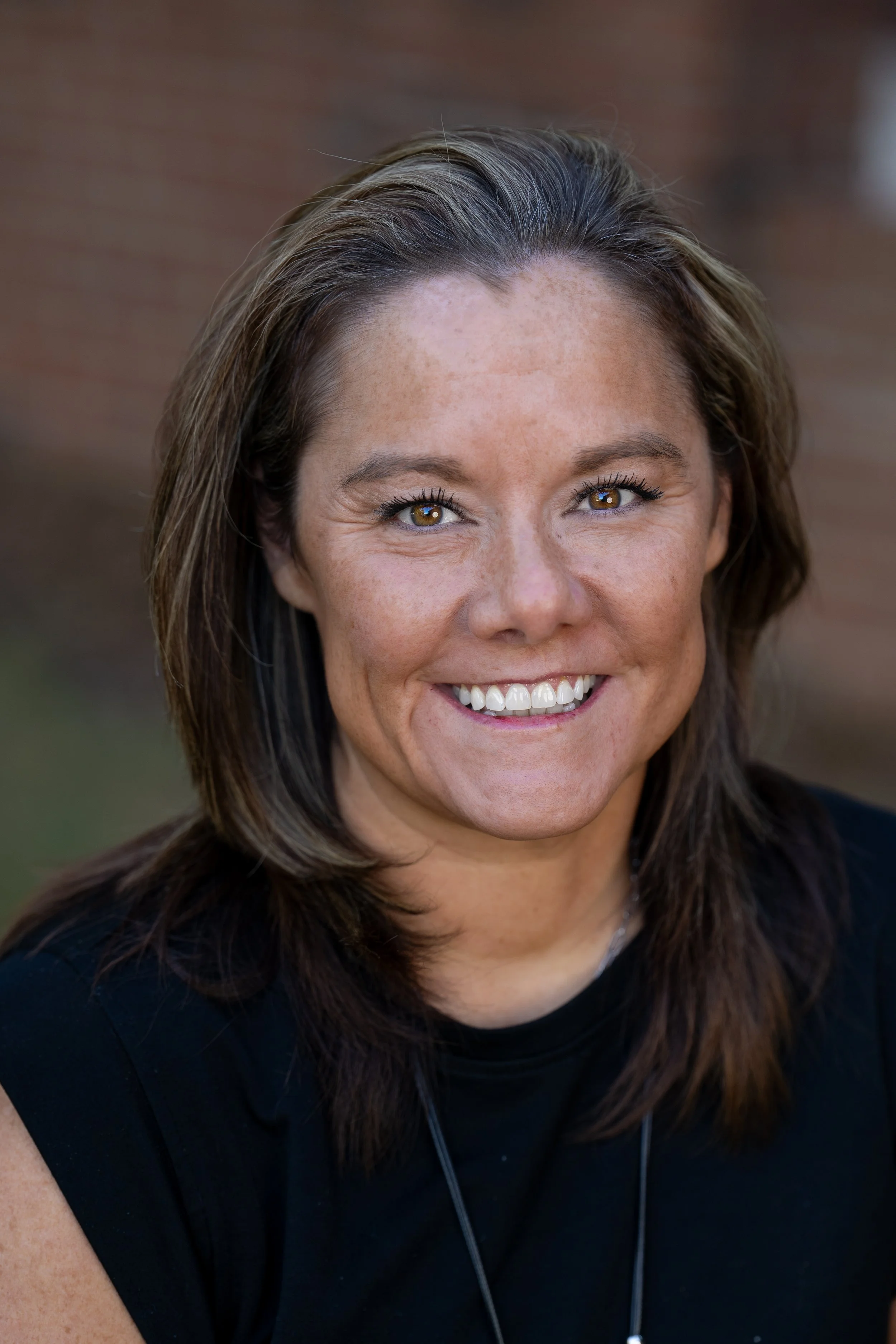 Close-up of a woman with shoulder-length brown hair smiling in front of a brick wall.