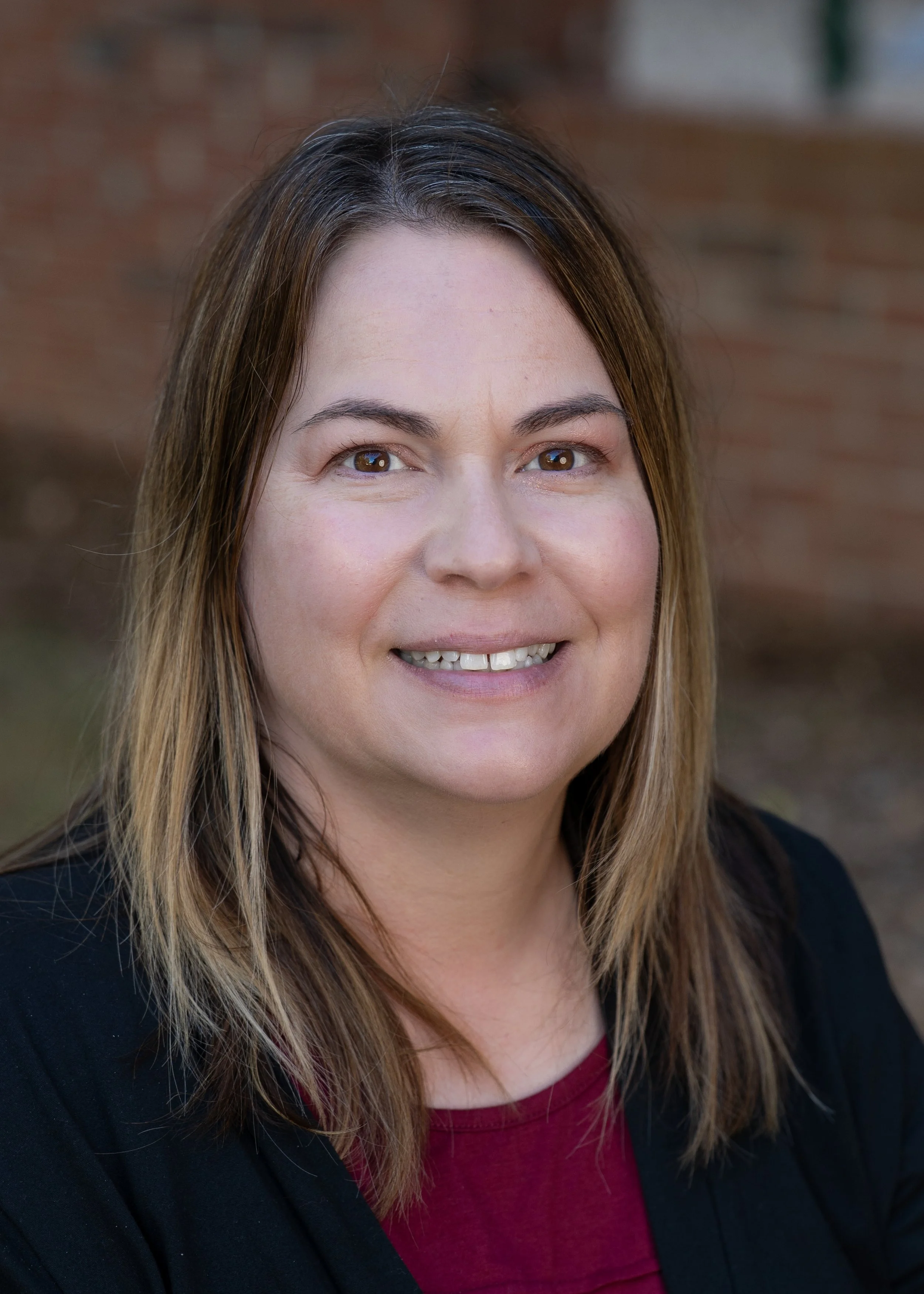 A woman with shoulder-length light brown hair standing in front of a brick wall.