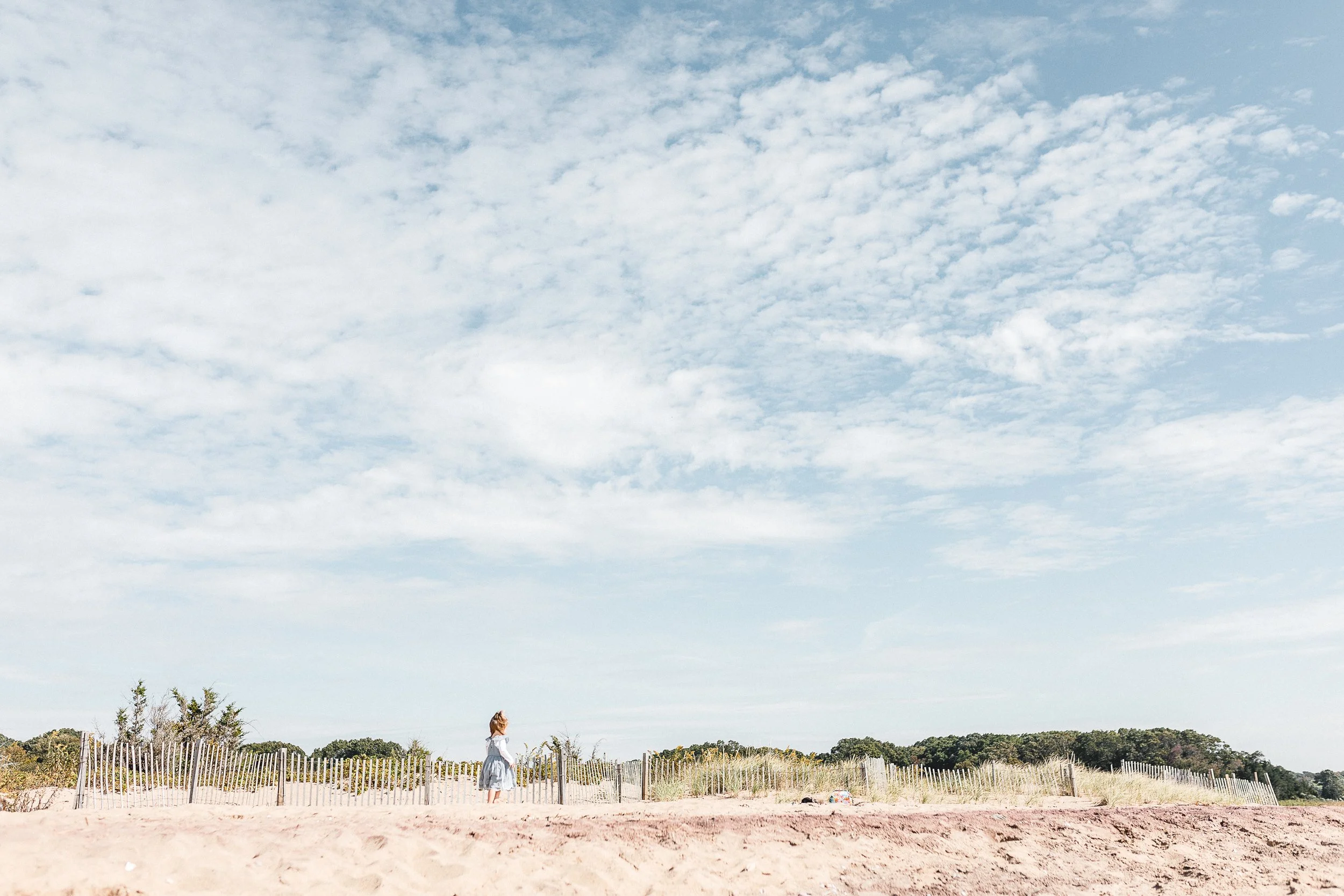 A person standing on a sandy beach near a wooden fence, with greenery in the background and a partly cloudy sky overhead.