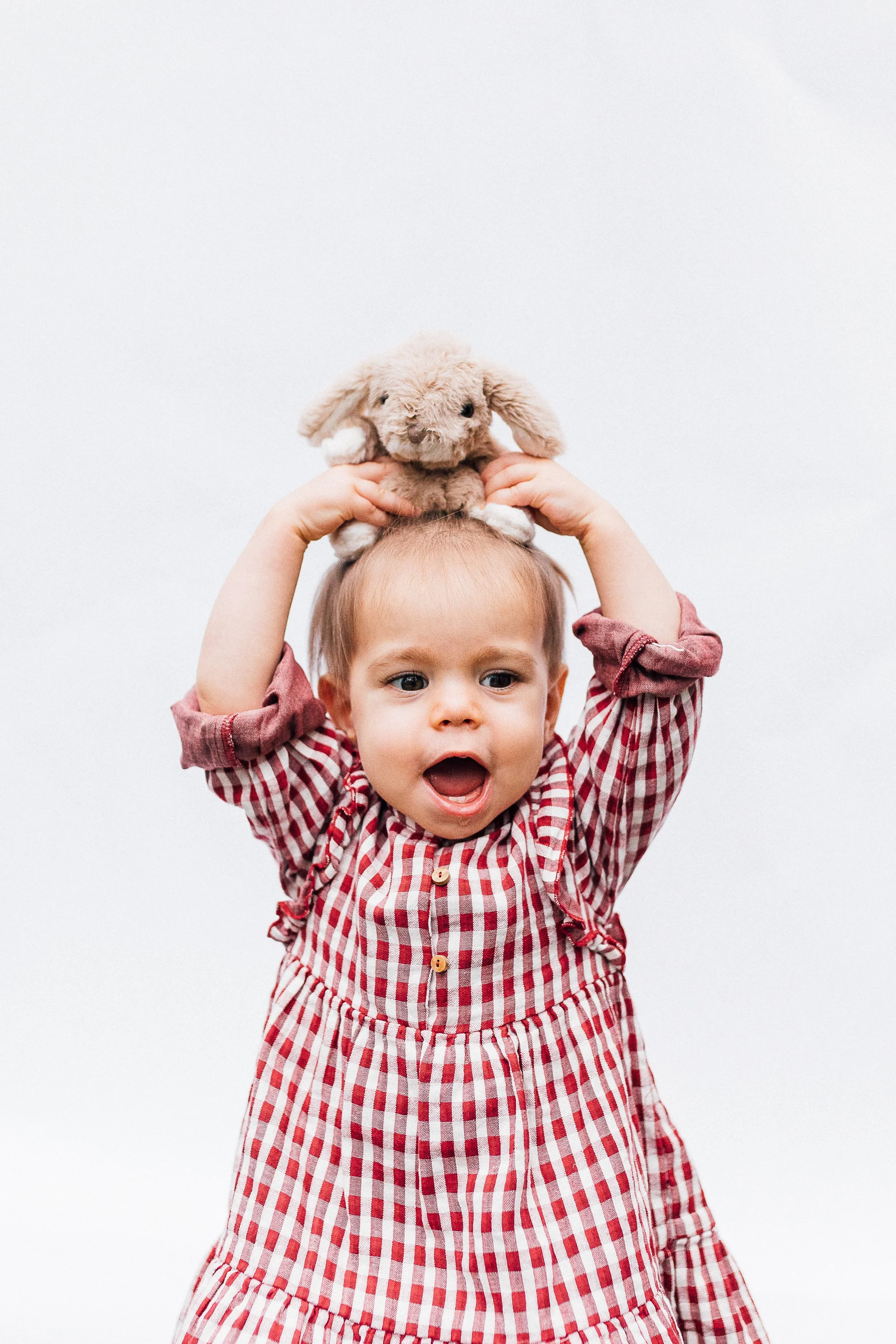 Young child in a red and white checkered dress holding a stuffed animal bunny on their head against a plain white background.