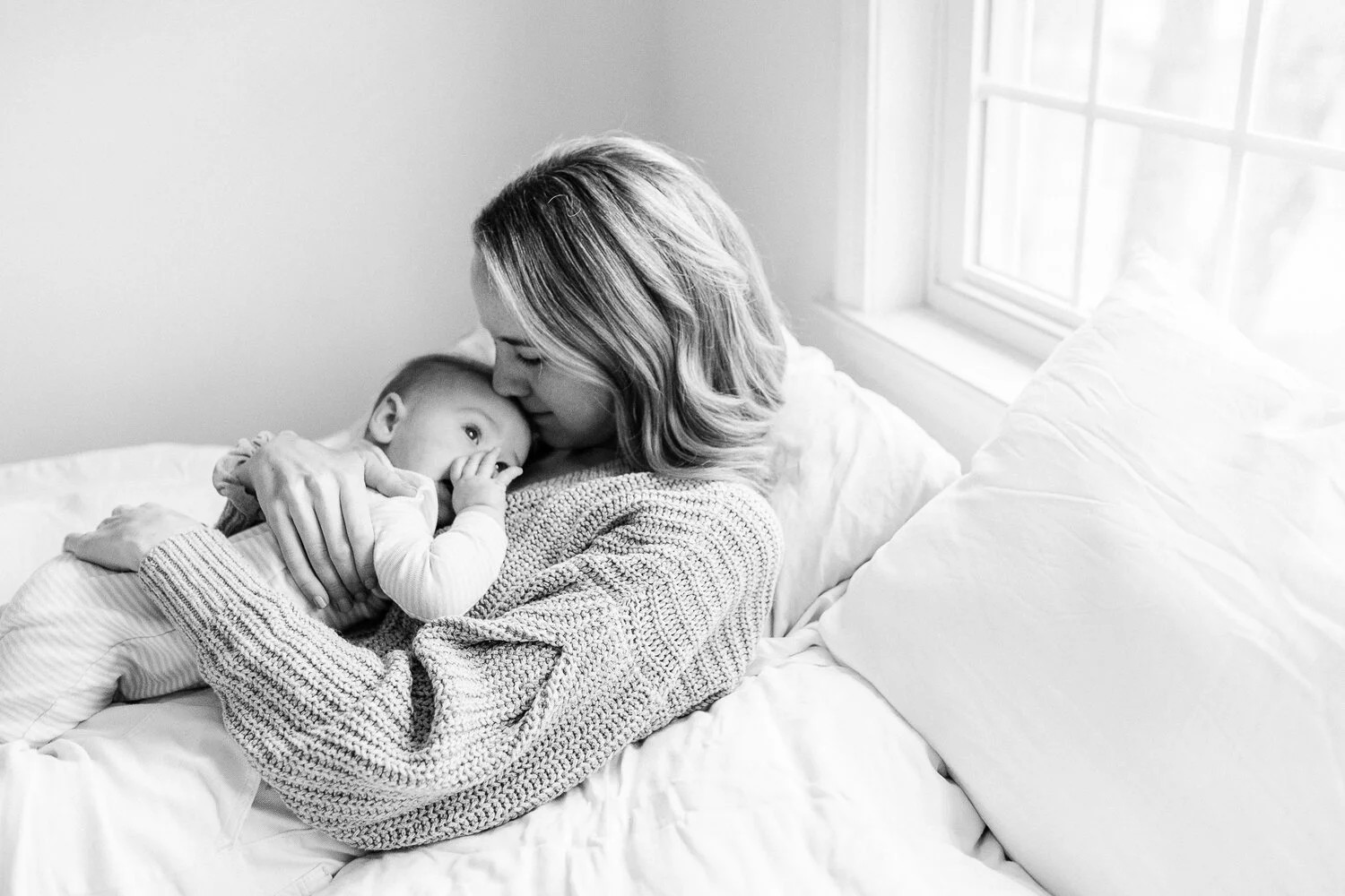 A woman cuddling a young child on a bed in a bright room with a window in the background.