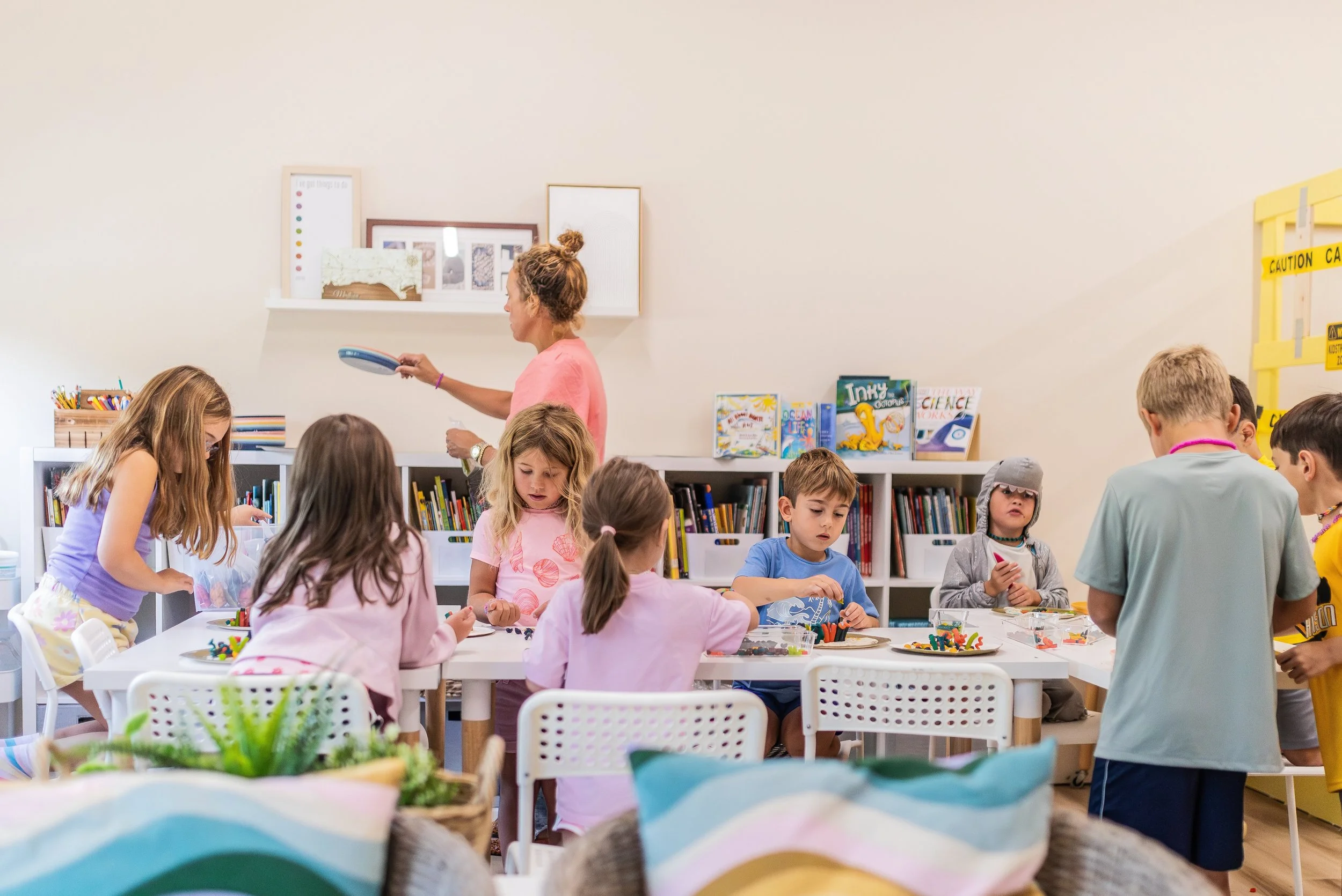 Children participating in a classroom activity with a teacher, sitting at tables with art supplies, bookshelves with books in the background, and hallway wall with yellow caution tape.