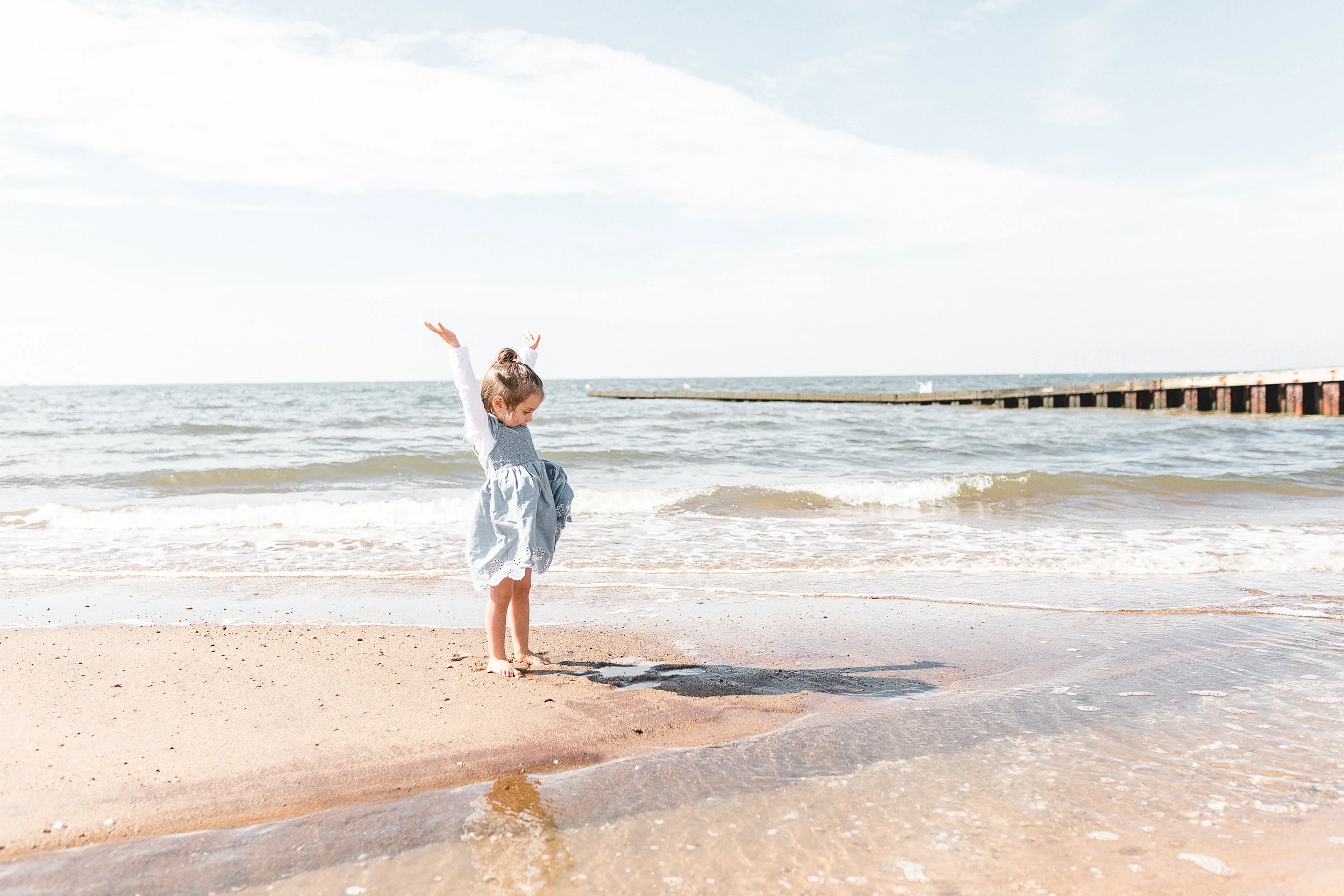 A young girl in a light blue dress and white bow in her hair playing on a sandy beach near the shoreline with waves and a wooden pier in the background.