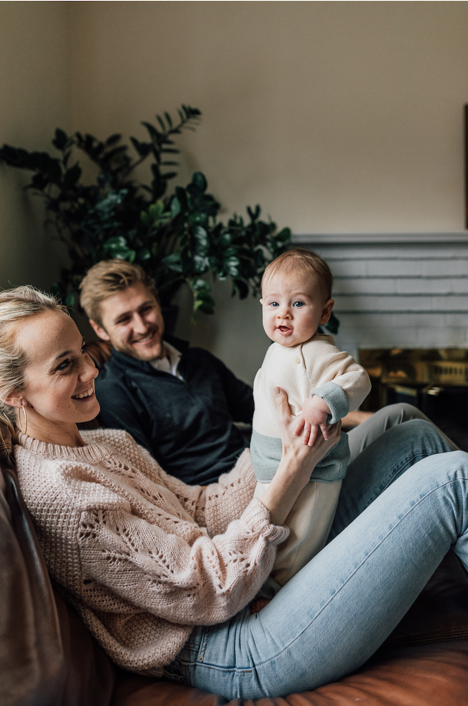 A happy woman holding a baby girl on her lap, with a smiling man sitting beside them on a couch in a cozy home setting, with a plant and fireplace in the background.