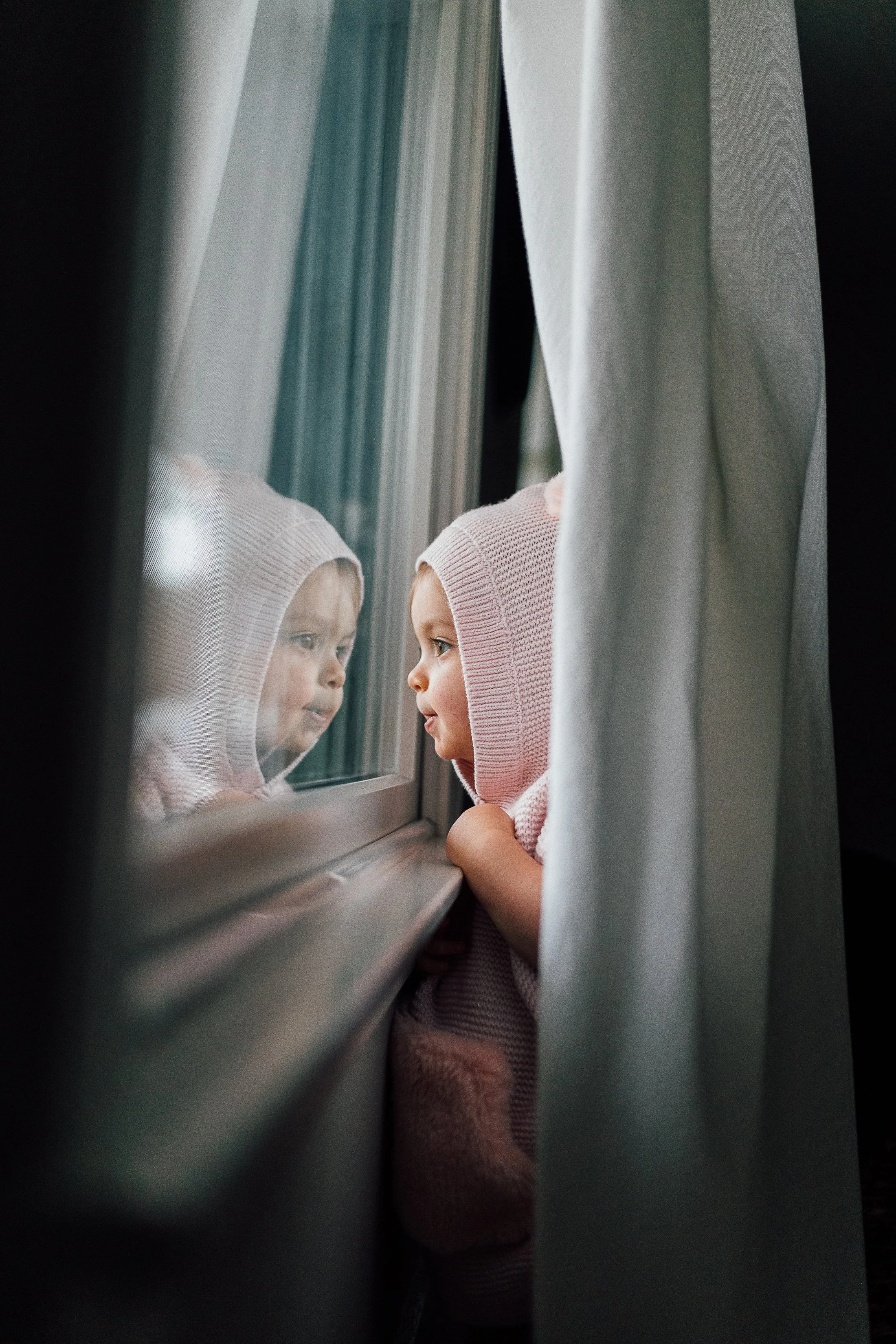 A young girl in a pink knit hoodie looking through a window, her reflection visible on the glass. She is leaning on the window ledge, partially covered by a curtain.
