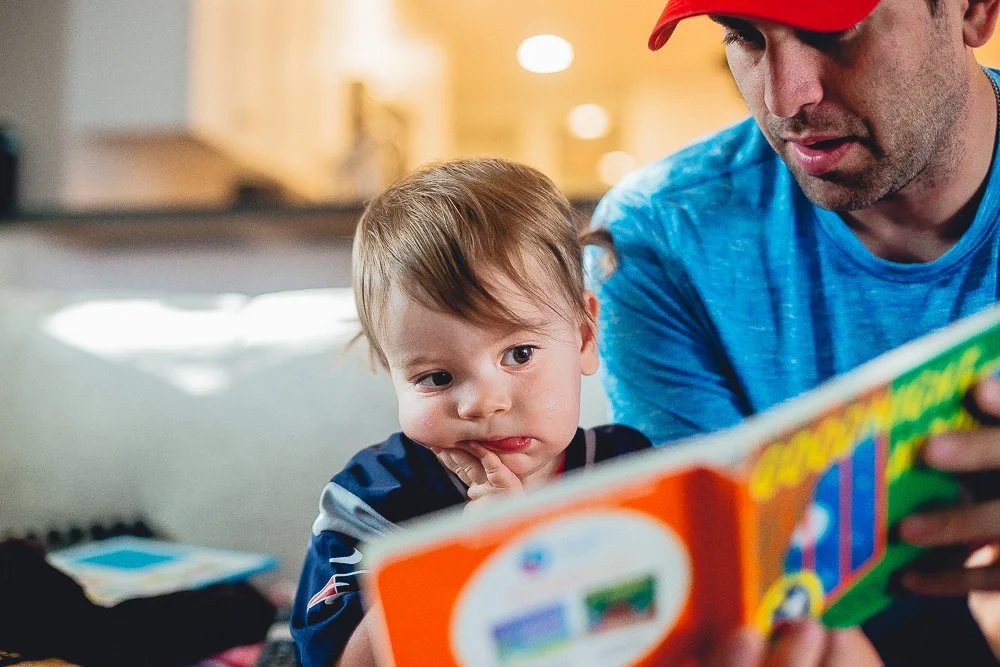A man and a young boy sit together, looking at a colorful toy or book. The boy appears curious or thoughtful, resting his chin on his hand, while the man is focused on the item they are holding. The background suggests a cozy indoor setting.