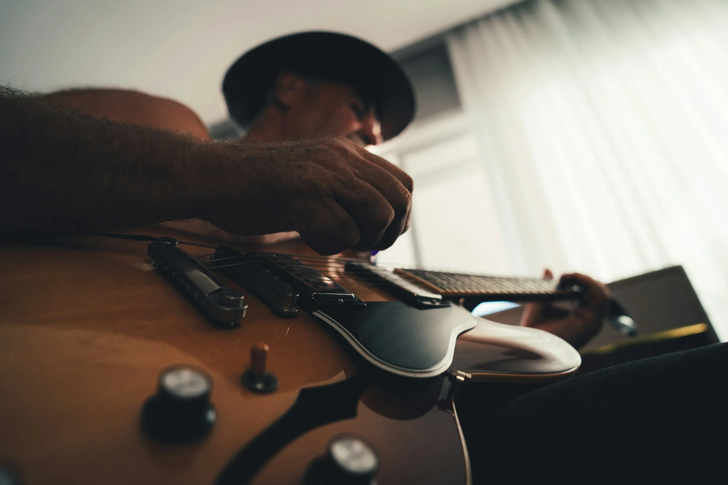 A person wearing a hat playing an electric guitar in a room with natural light coming through a window with curtains.