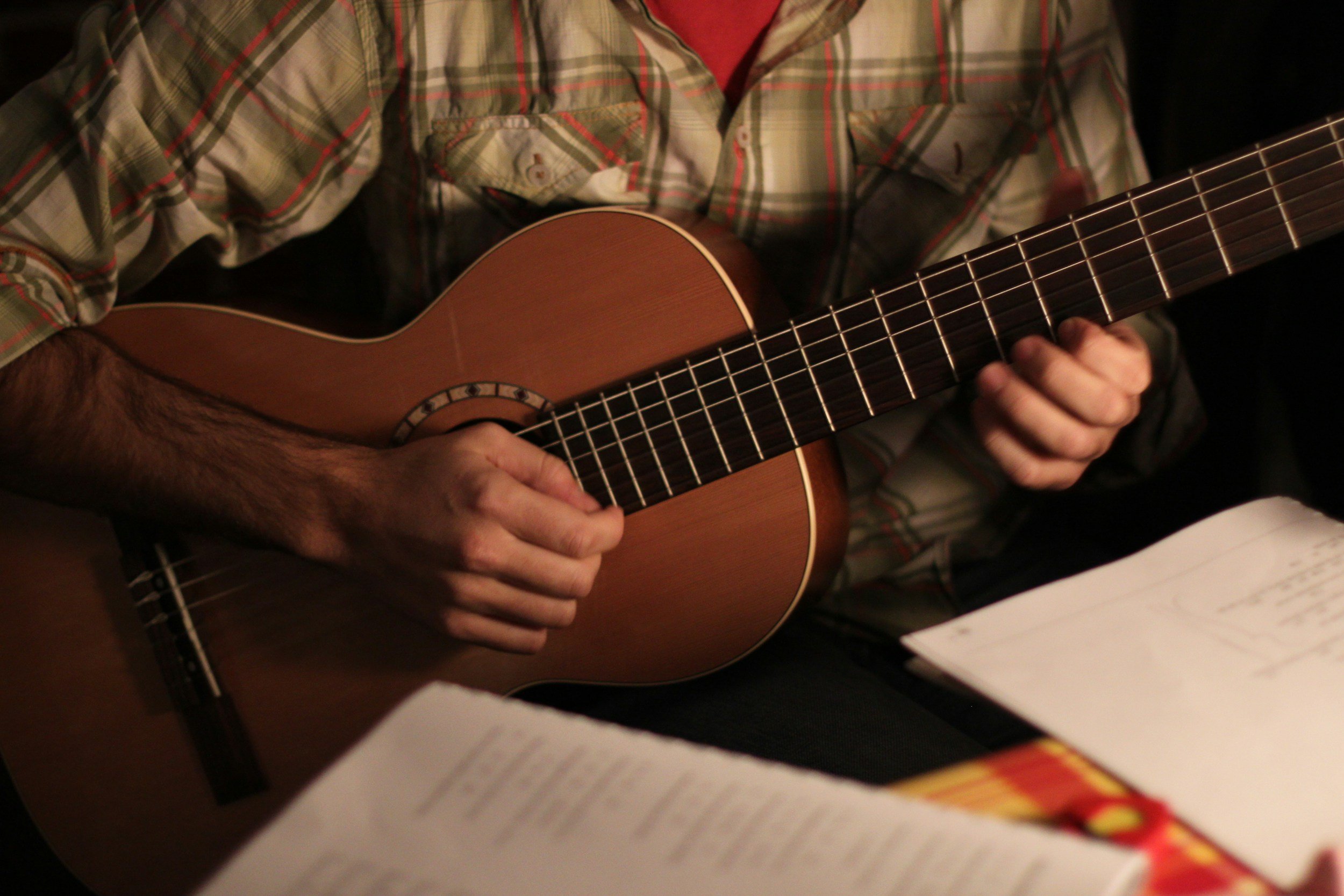 A person playing an acoustic guitar, wearing a plaid shirt, with sheet music or lyrics visible in front of them.