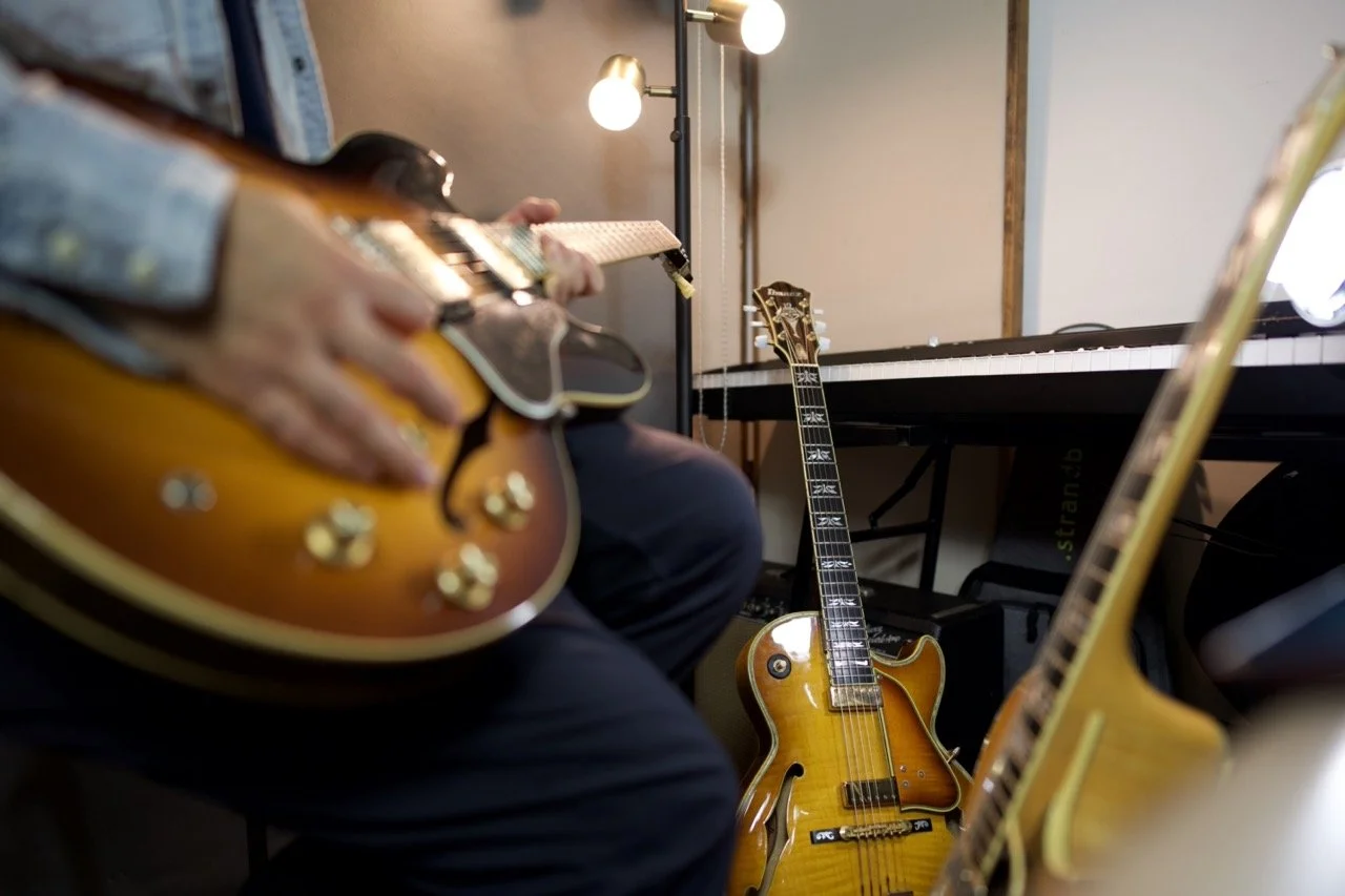 Person playing an electric guitar in a music studio, with a second electric guitar resting against a keyboard in the background.
