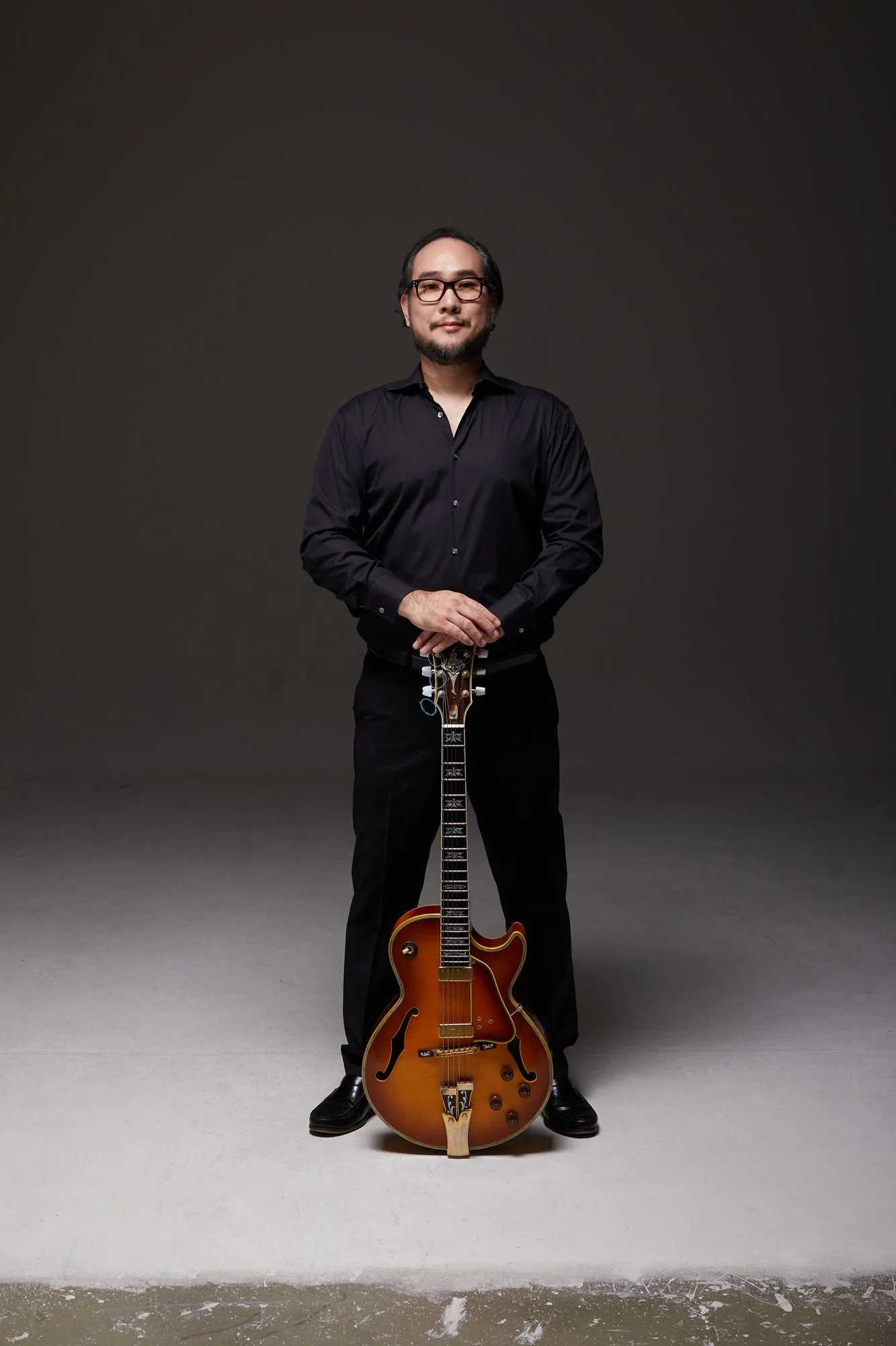 A man with glasses, a beard, and shoulder-length black hair stands against a dark gray background, holding a vintage orange semi-hollow electric guitar with f-holes.