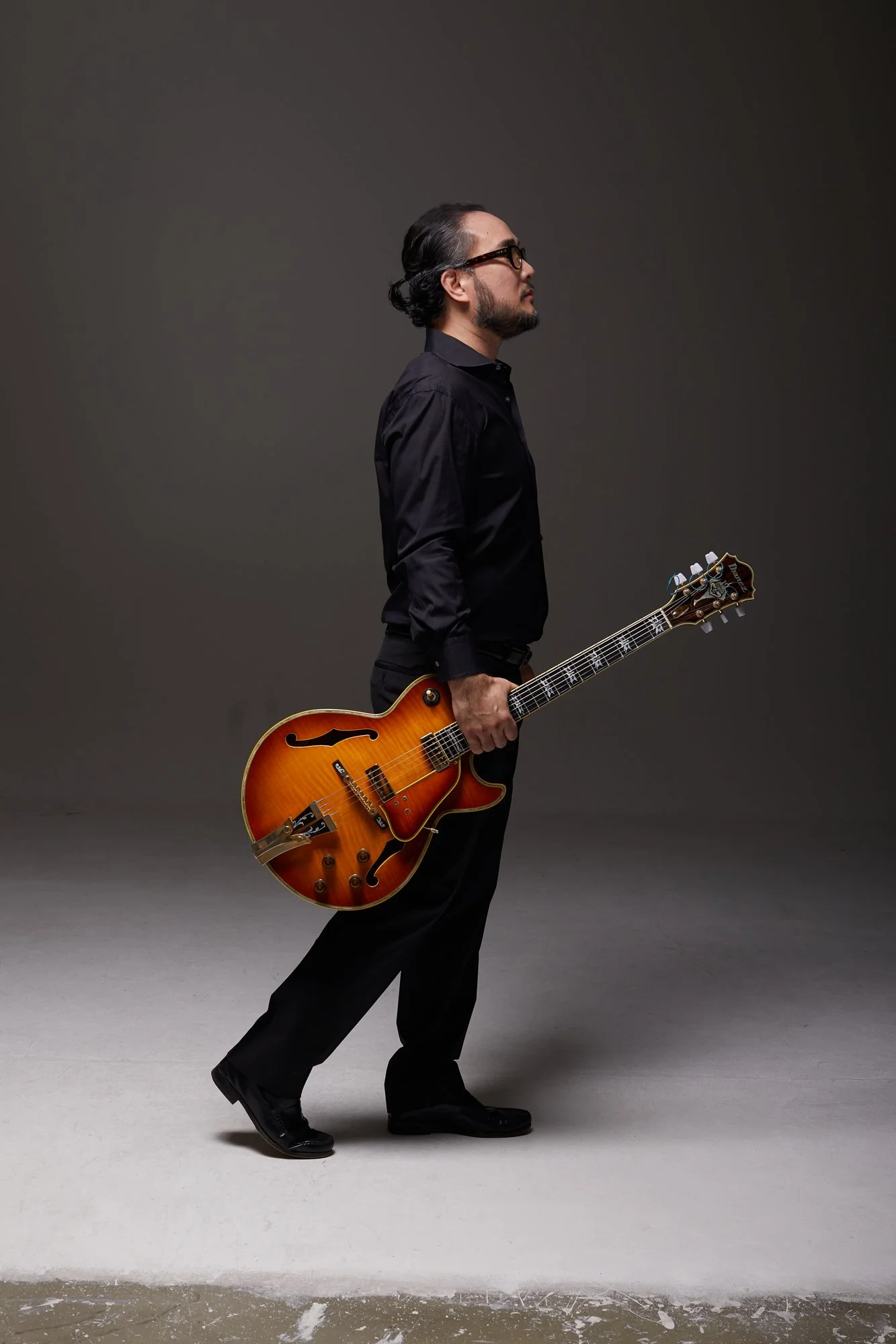 A man in black clothing and glasses is standing with his eyes closed, holding a semi-hollow body electric guitar, in a studio with a dark gray background and a light gray floor.