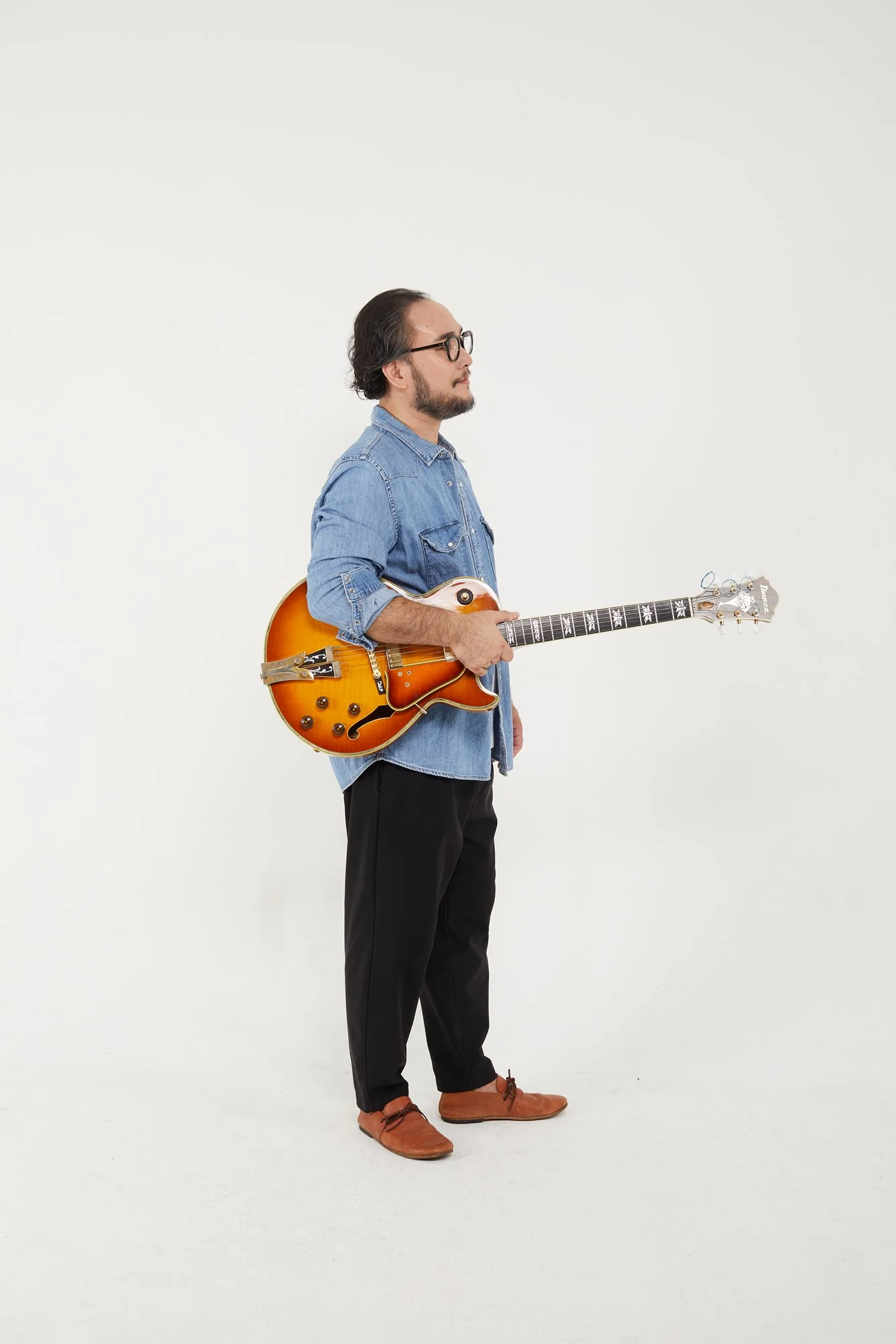 A man with glasses, a beard, and long hair tied back, dressed in a denim shirt and black pants, standing sideways, holding a vintage orange sunburst electric guitar.