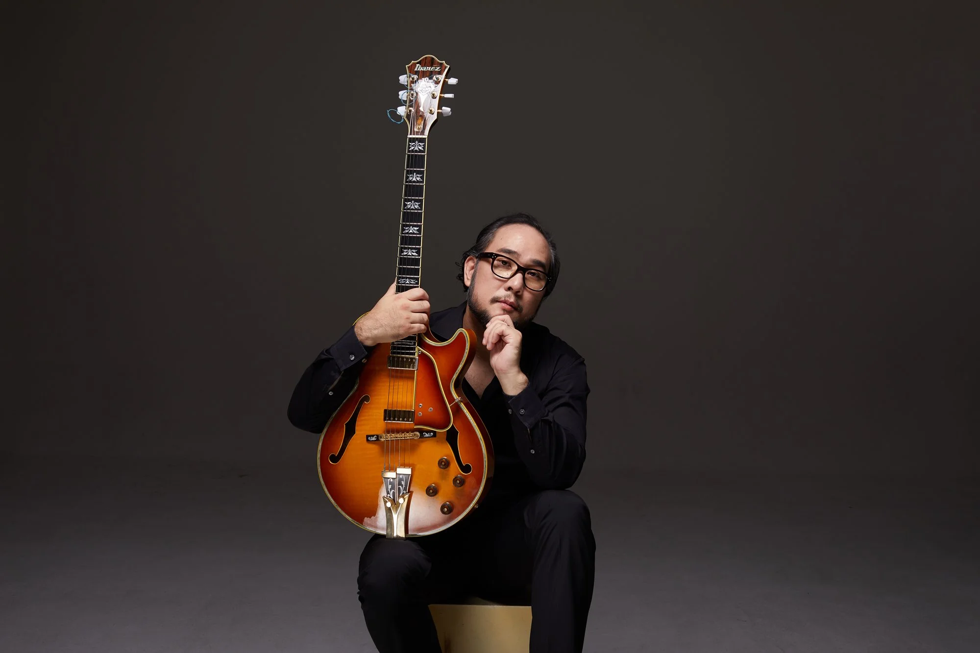 A man with glasses and a beard sitting on a yellow stool, holding a vintage orange electric guitar with a semi-hollow body design against a dark background.