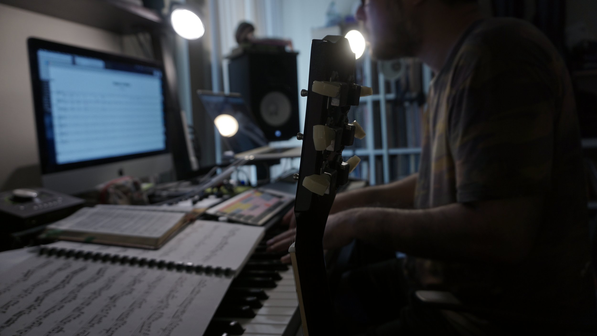 A person playing an acoustic guitar, wearing a plaid shirt, with sheet music or lyrics visible in front of them.